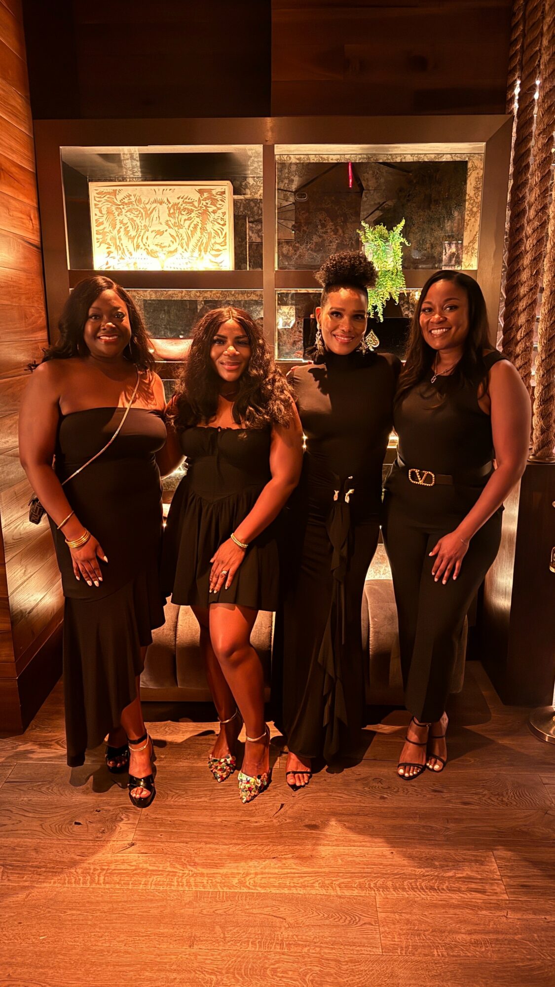 Four women standing together indoors, smiling, with a window and curtains behind them.