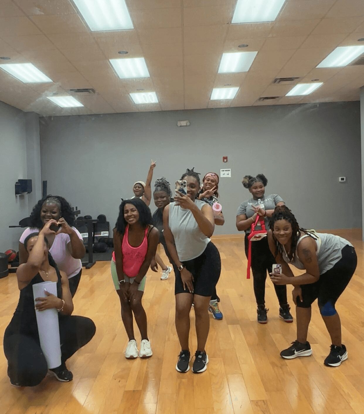 Group of women and girls in a dance studio, posing and smiling, some making peace signs, with a wooden floor and ceiling lights.