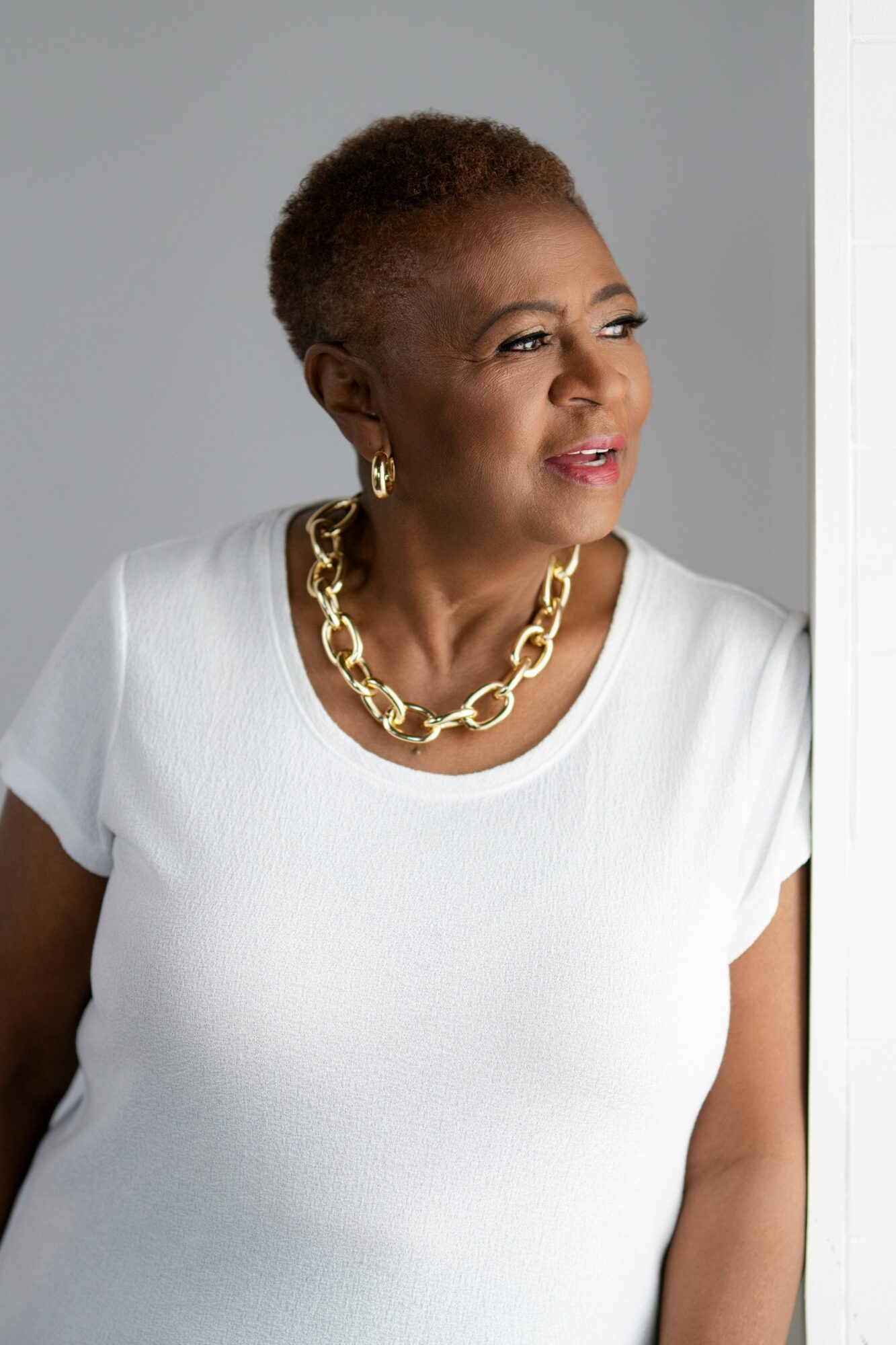 Woman with short hair wearing a white top, gold chain necklace, and earrings, looking out a window.