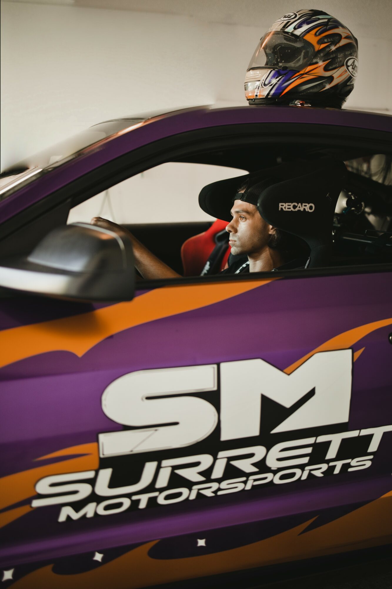 Person in racing helmet sitting in a purple race car with 'SM Surrett Motorsports' logo, driver wearing a black cap, inside a garage.
