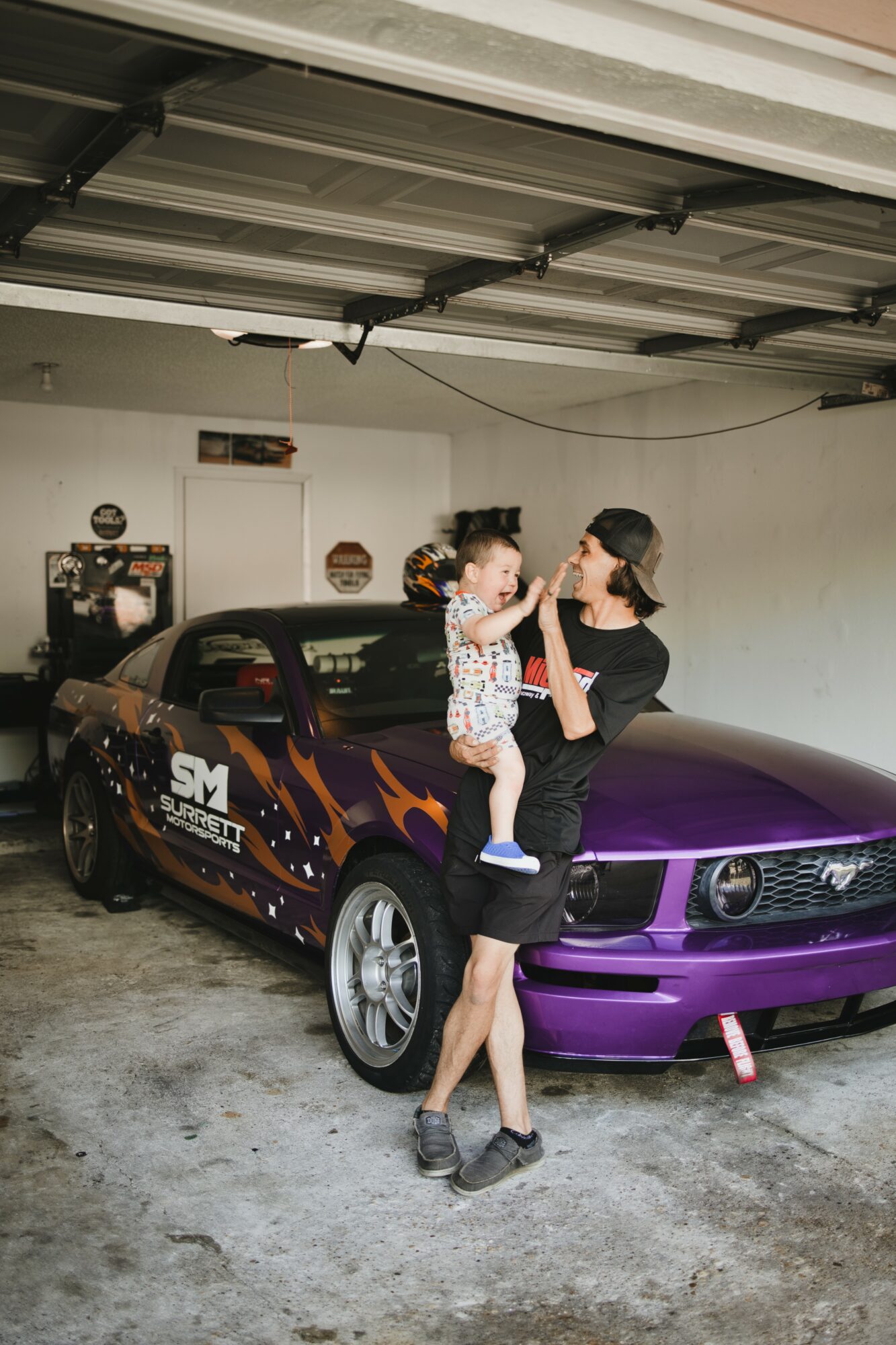 Woman holding a child in front of a purple and black sports car inside a garage.
