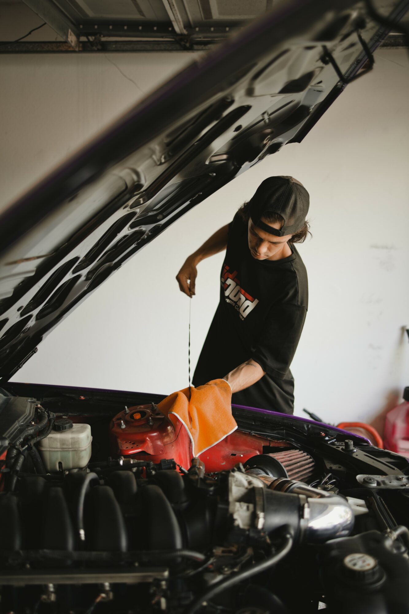 Person inspecting car engine with hood open, wearing a black cap and shirt, in a garage setting.
