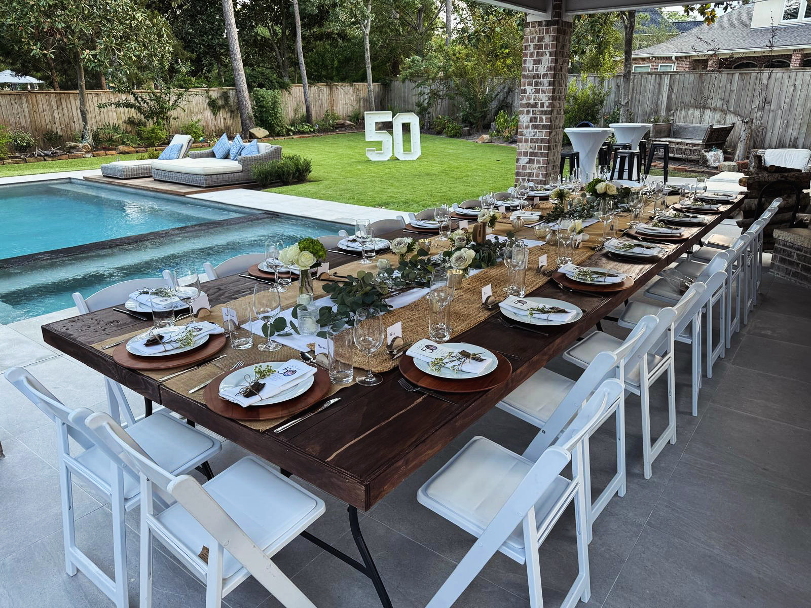 Long outdoor dining table set with plates, glasses, and cutlery, overlooking a backyard with a pool and garden.