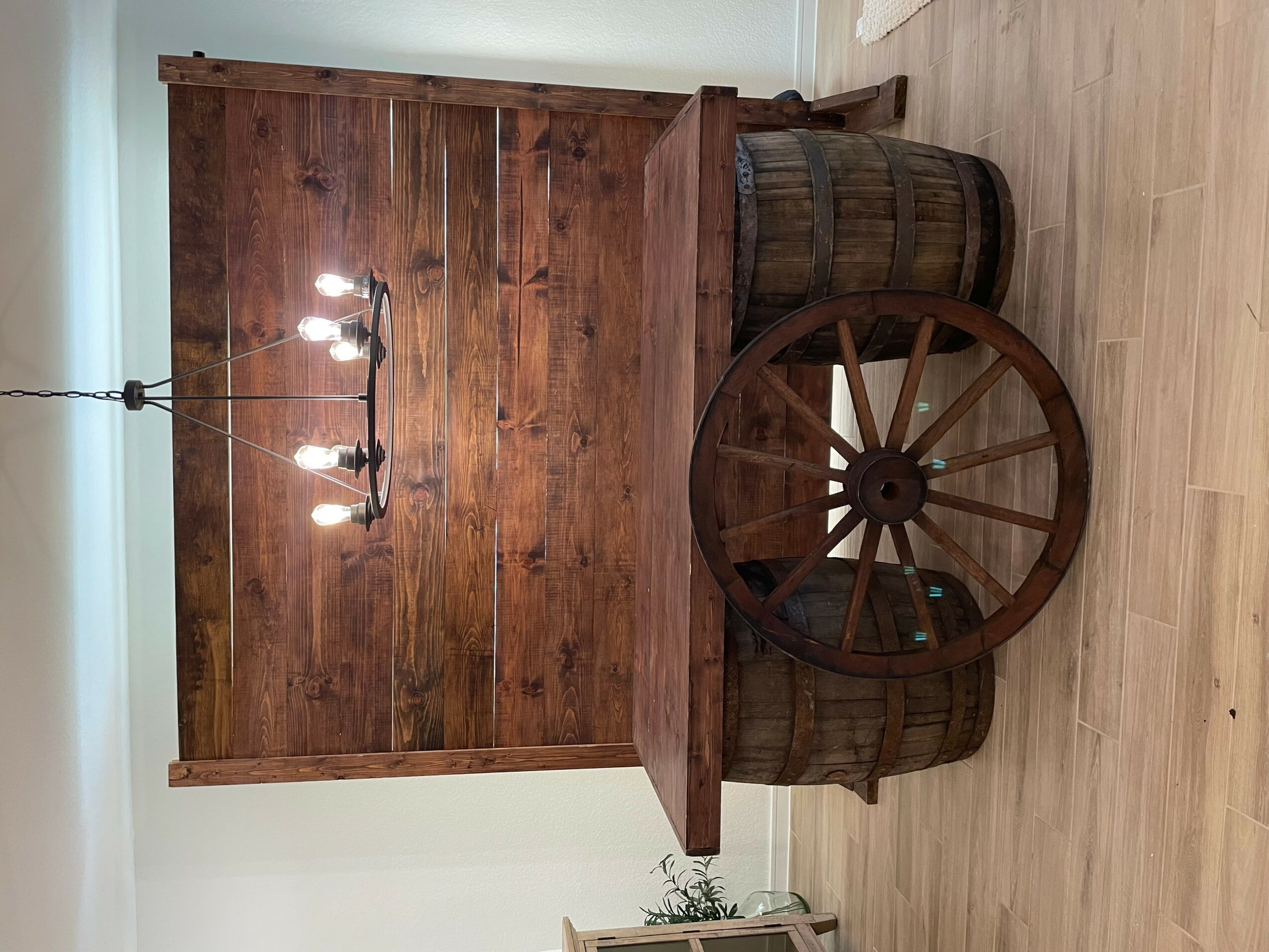 Wooden bar with barrel stools and a large wooden wheel, against a wooden wall, on a light-colored floor.
