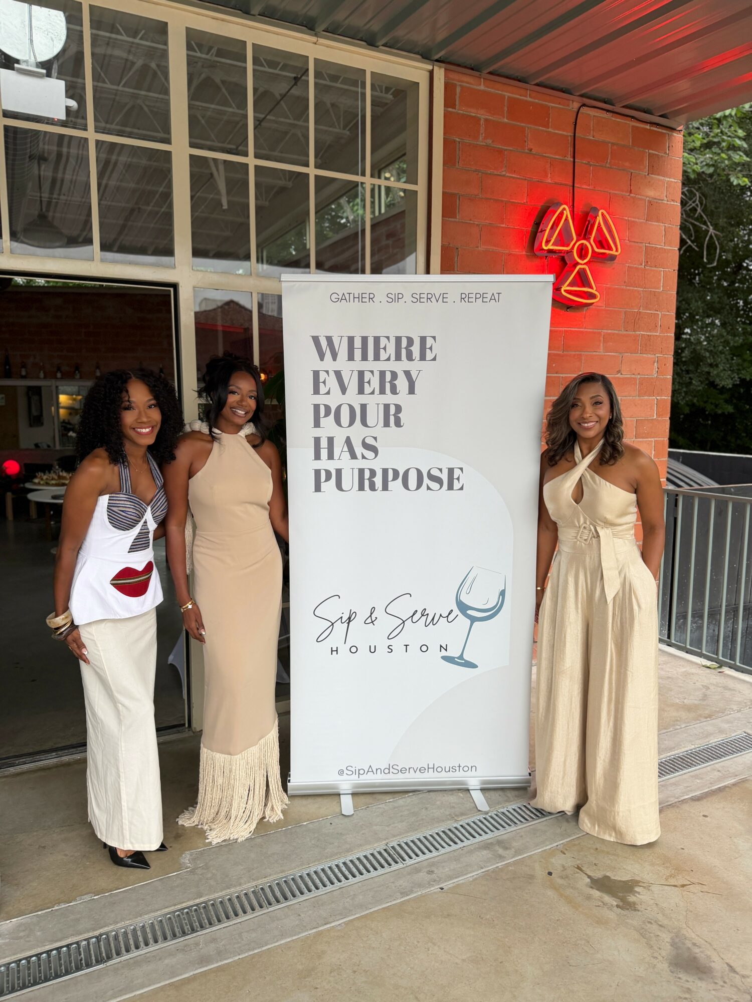 Three women in elegant dresses stand outside a building with a large sign and a red radiation symbol neon light.