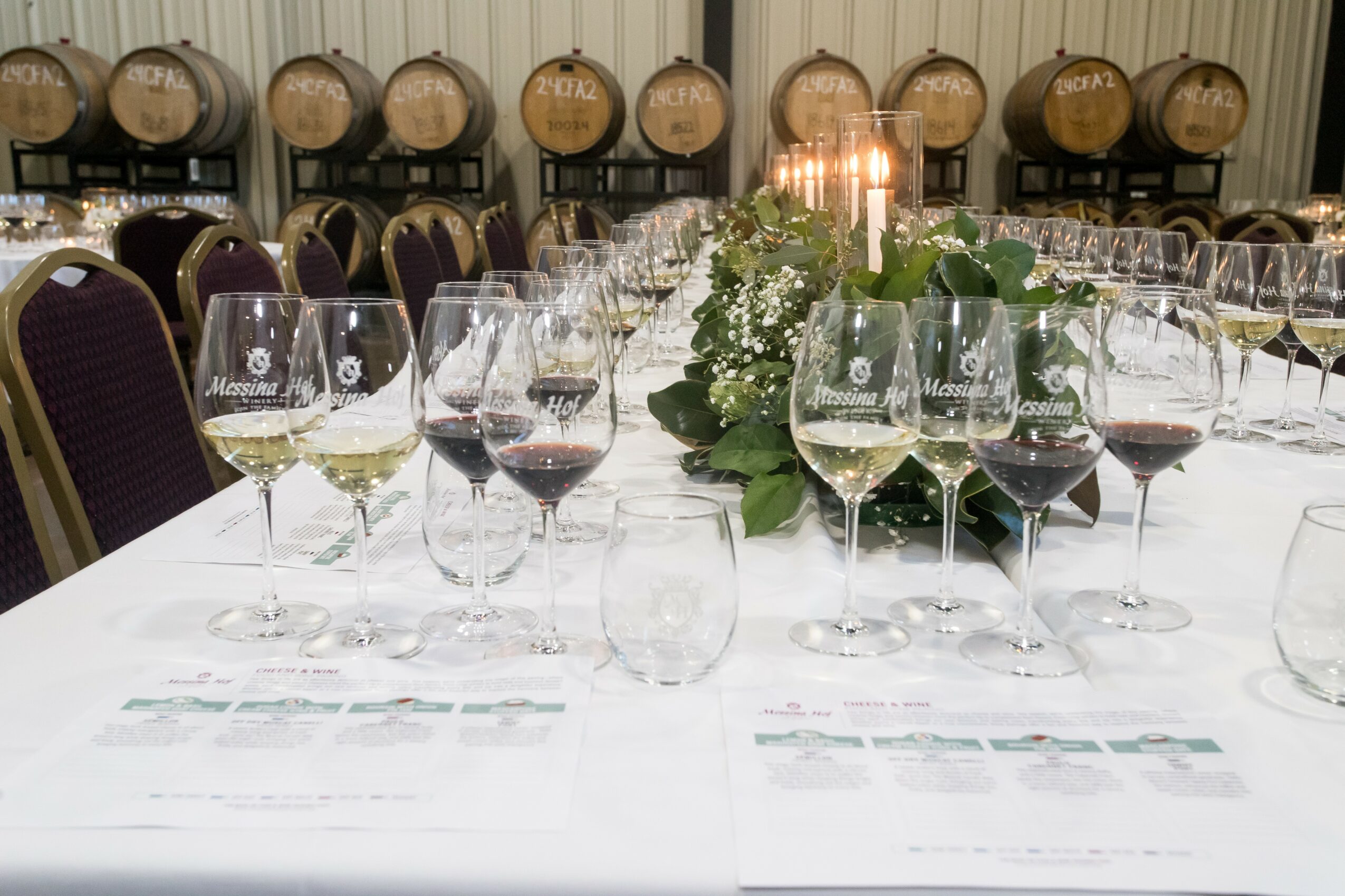 Long table set with wine glasses, a floral centerpiece, and wine barrels in the background, in a banquet hall.