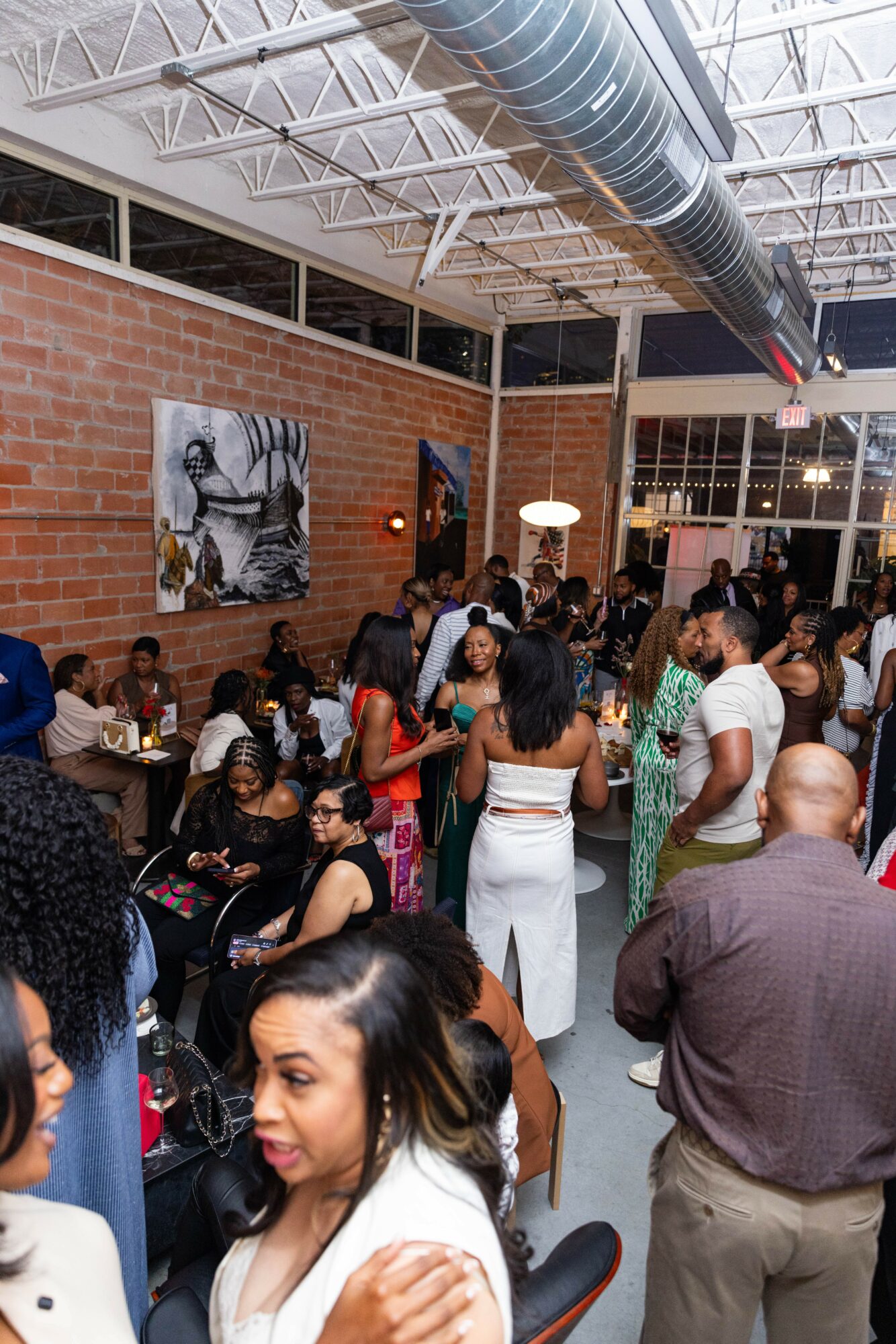 People gather in a spacious indoor venue with brick walls, artwork, and large windows, socializing and enjoying the event.