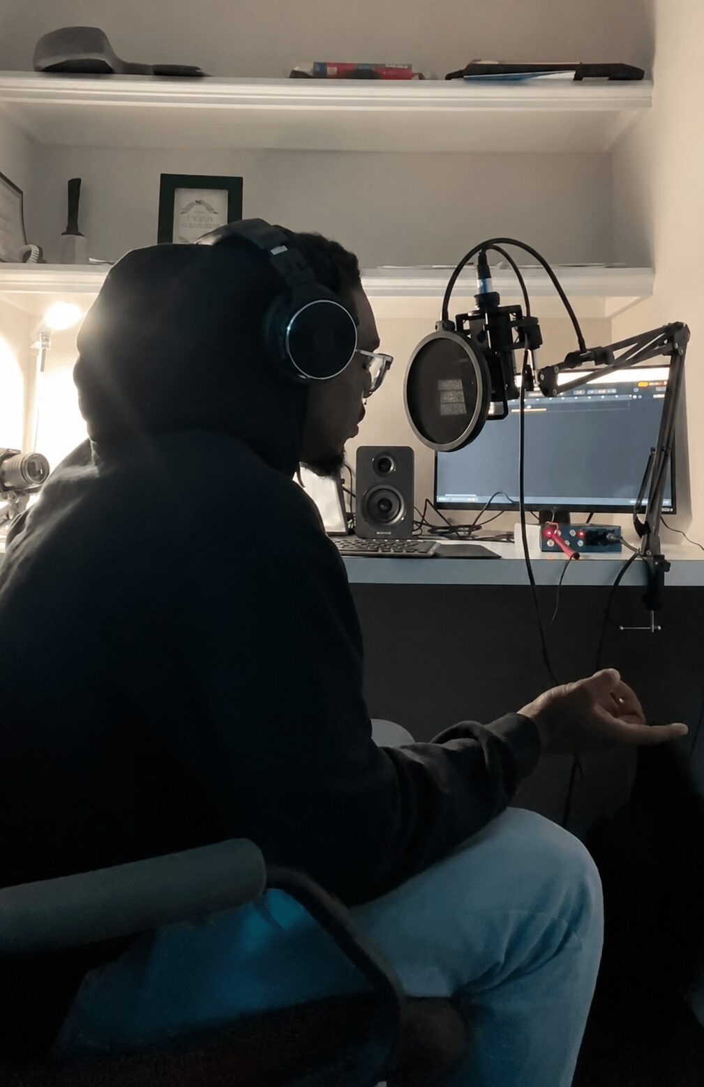 Person wearing headphones sitting at a desk with a microphone, computer, and audio equipment in a room.
