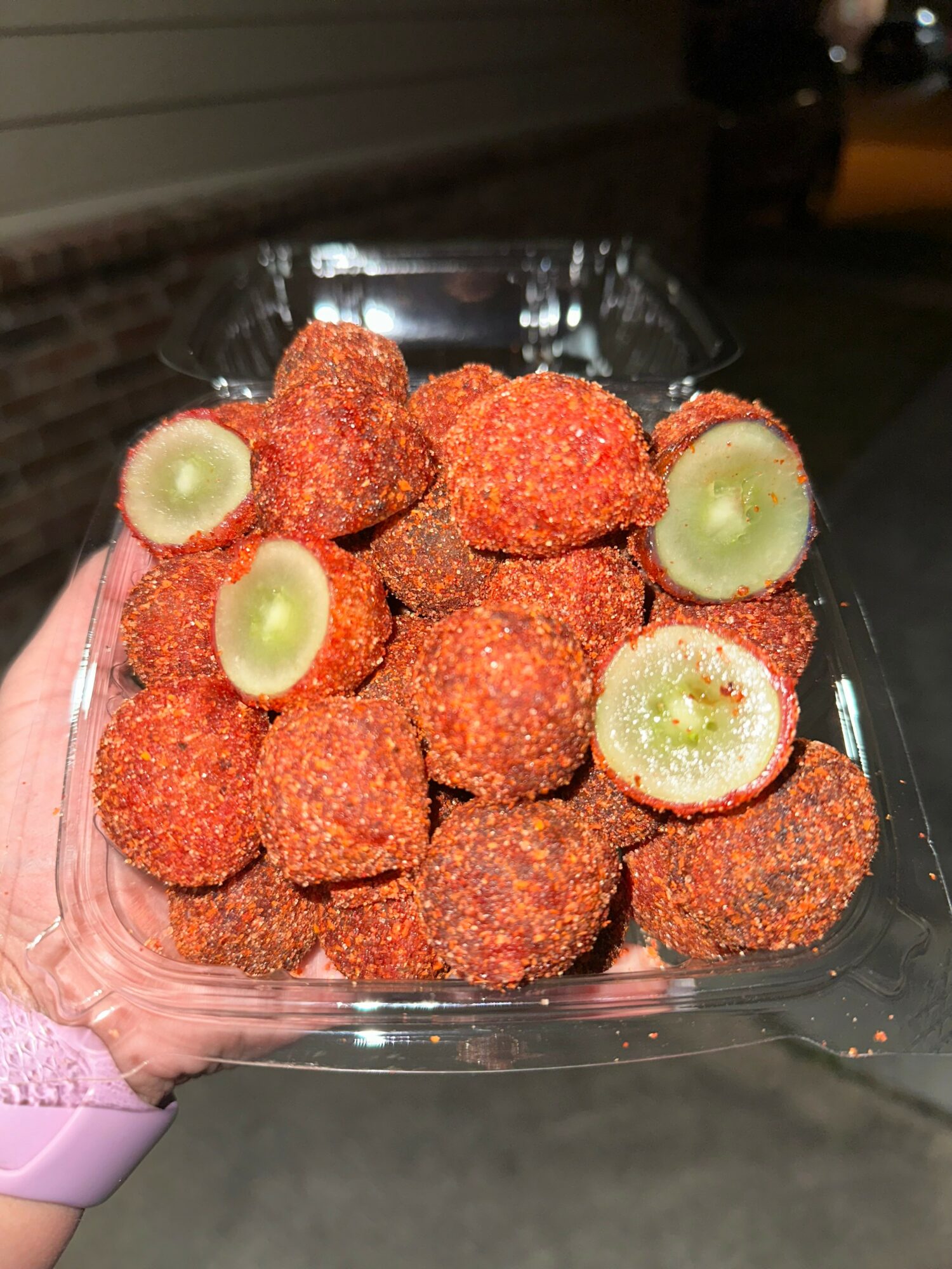 Hand holding a clear container with breaded, fried food and sliced green vegetables inside, outdoors at night.