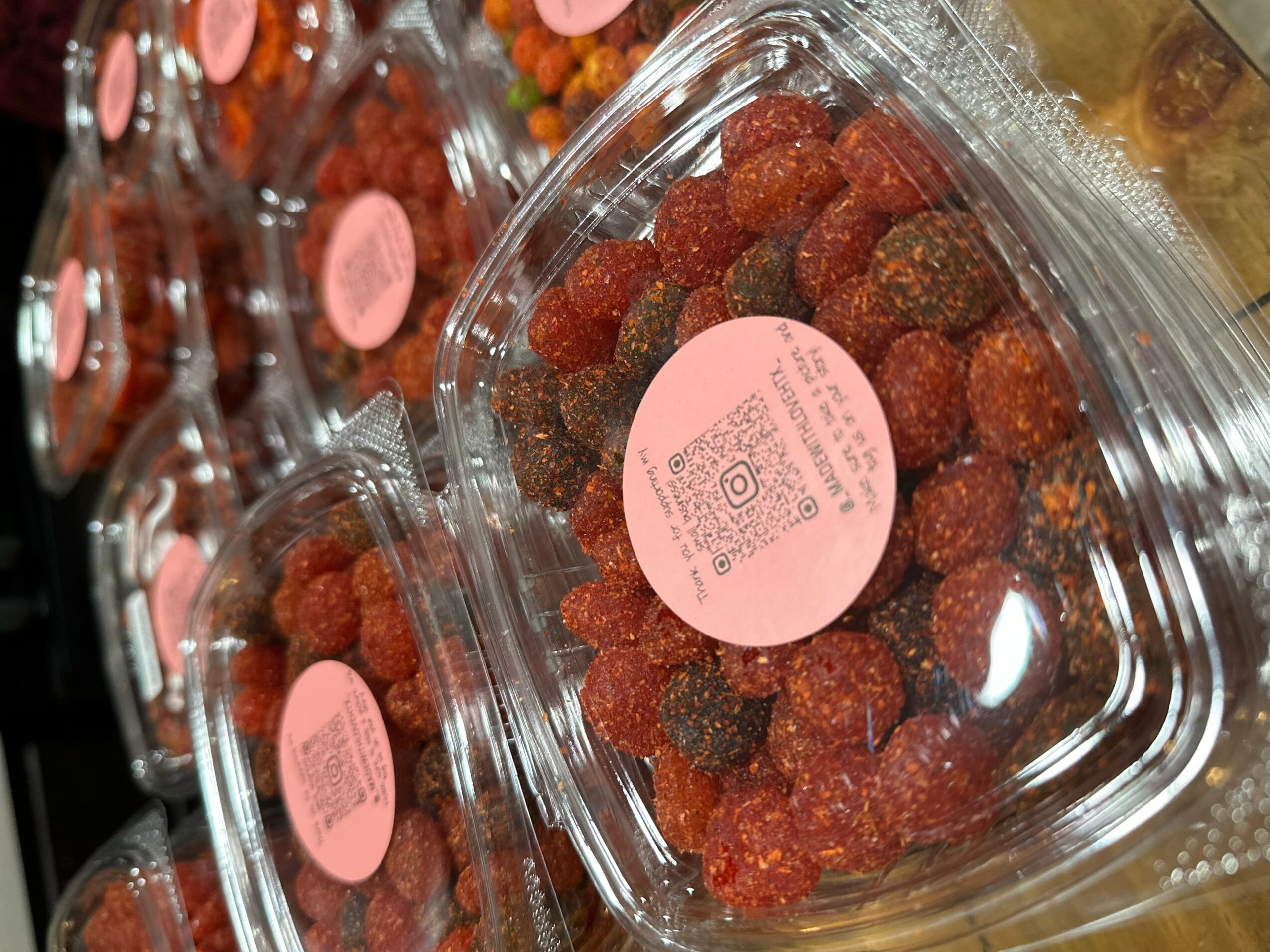 Clear plastic container filled with red and dark berries, with a pink label on top, on a shelf with similar containers