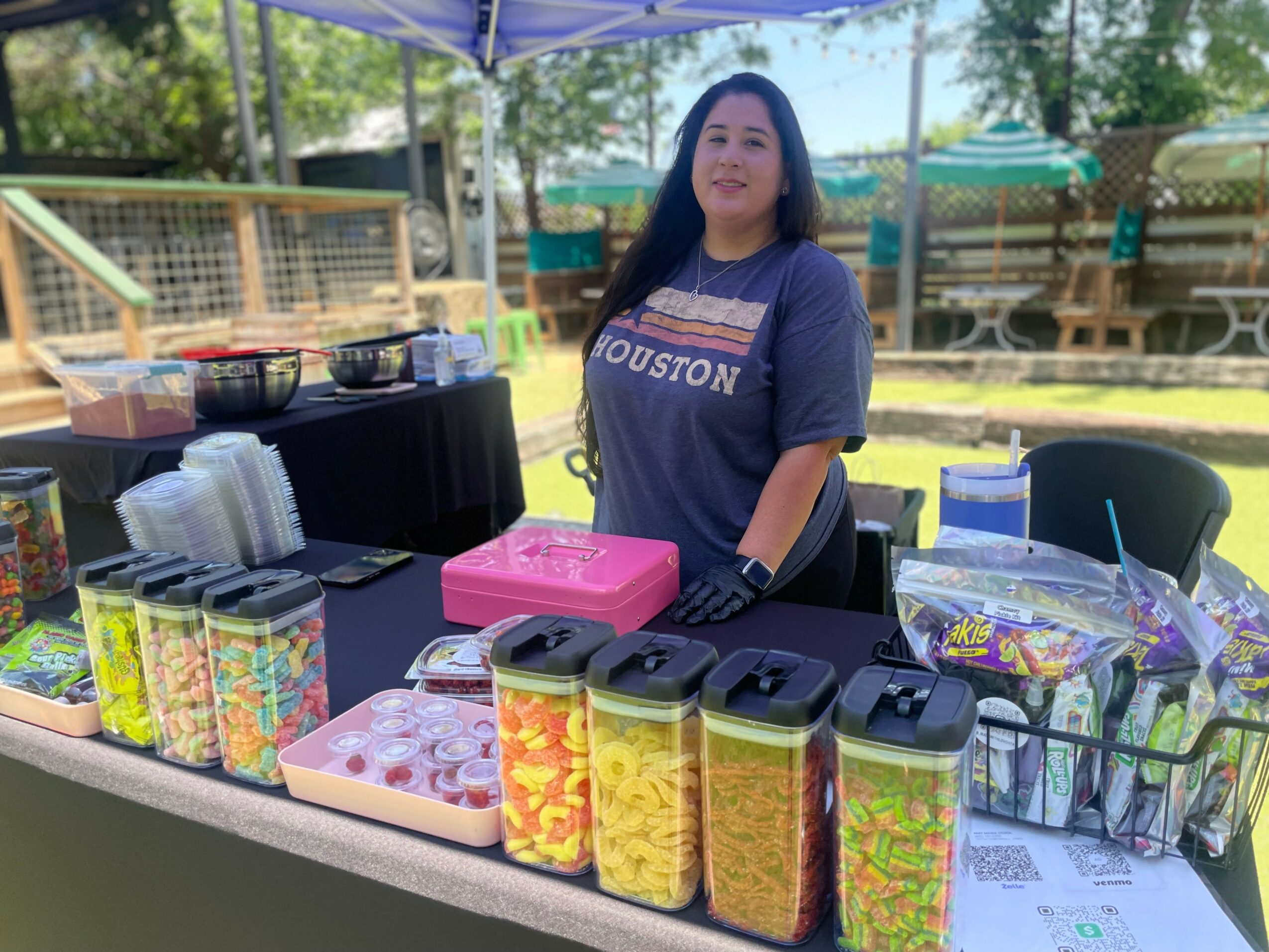 Young woman standing behind a table with colorful snacks and supplies outdoors, under a canopy, with trees and a fence in background.