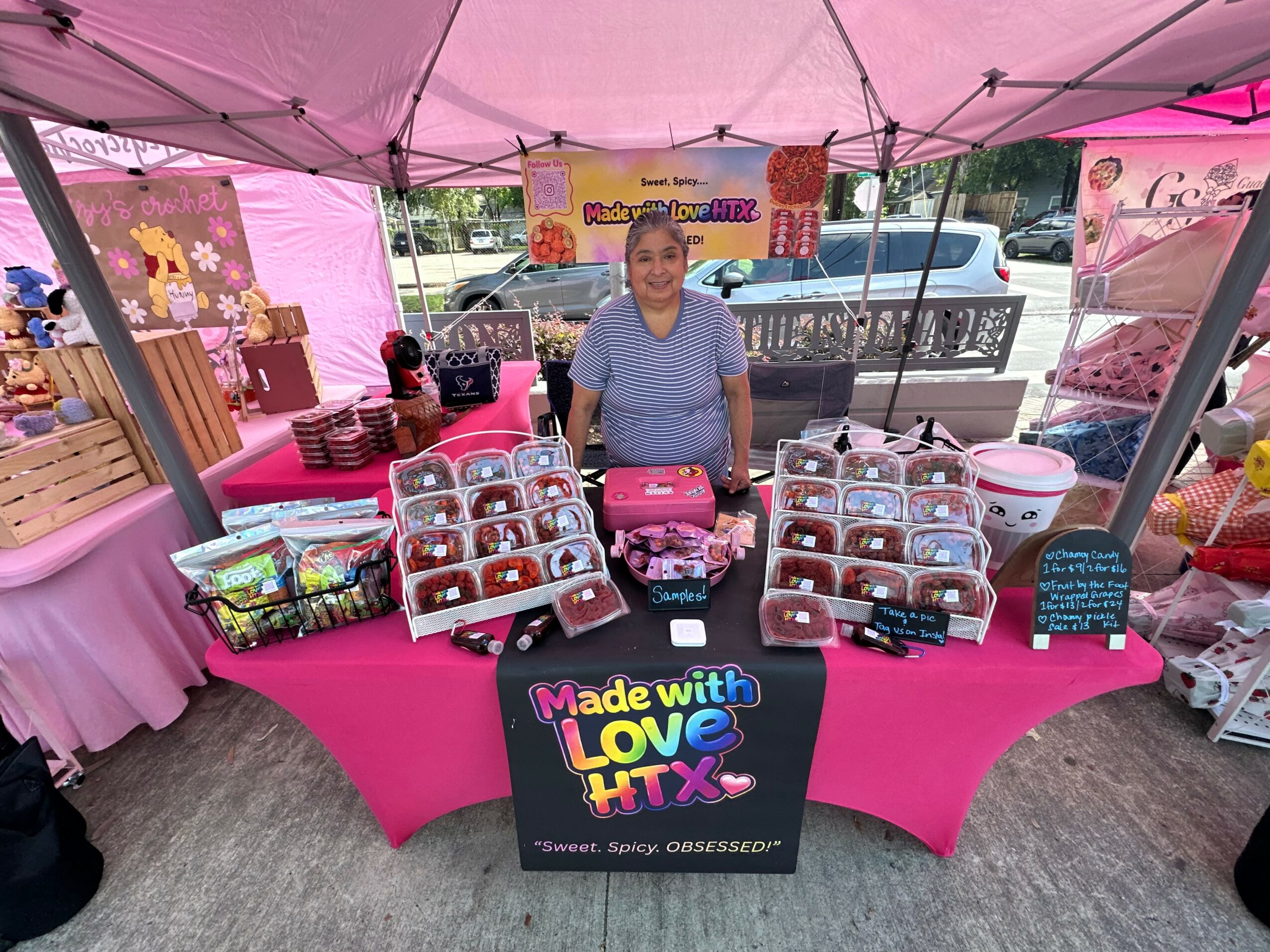 Woman standing behind a pink table with jars and baked goods at a market stall, pink canopy overhead, colorful signs, and other stalls nearby.