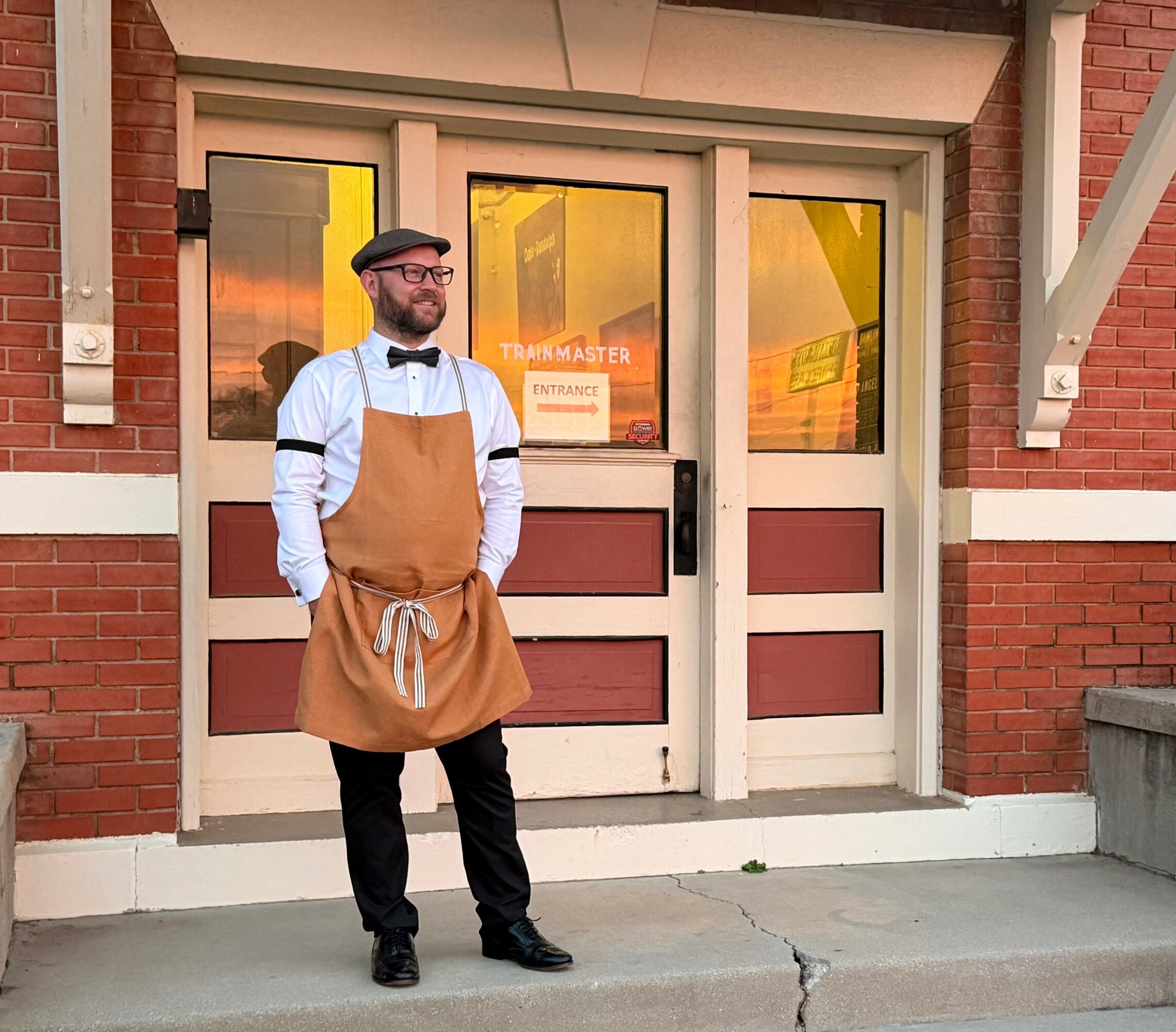 Man in apron standing outside building with brick walls and glass door, sunlight reflecting in windows.