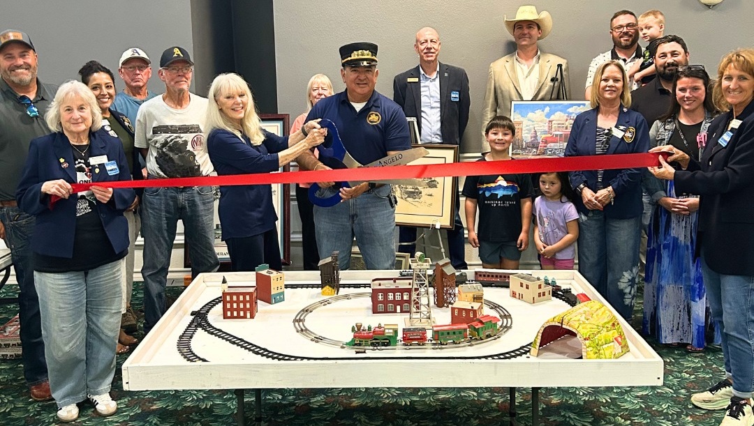 Group of people gathered around a model train display, with some holding a red ribbon for a ribbon-cutting event.