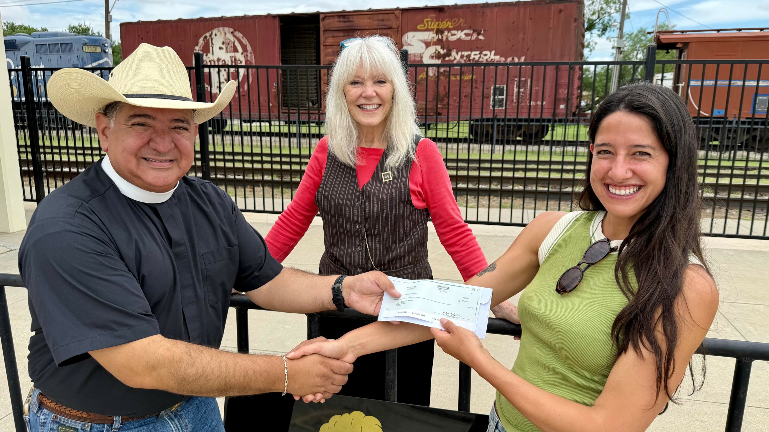 Three people outdoors, a man in cowboy hat and a woman in green shirt exchanging a check, smiling.