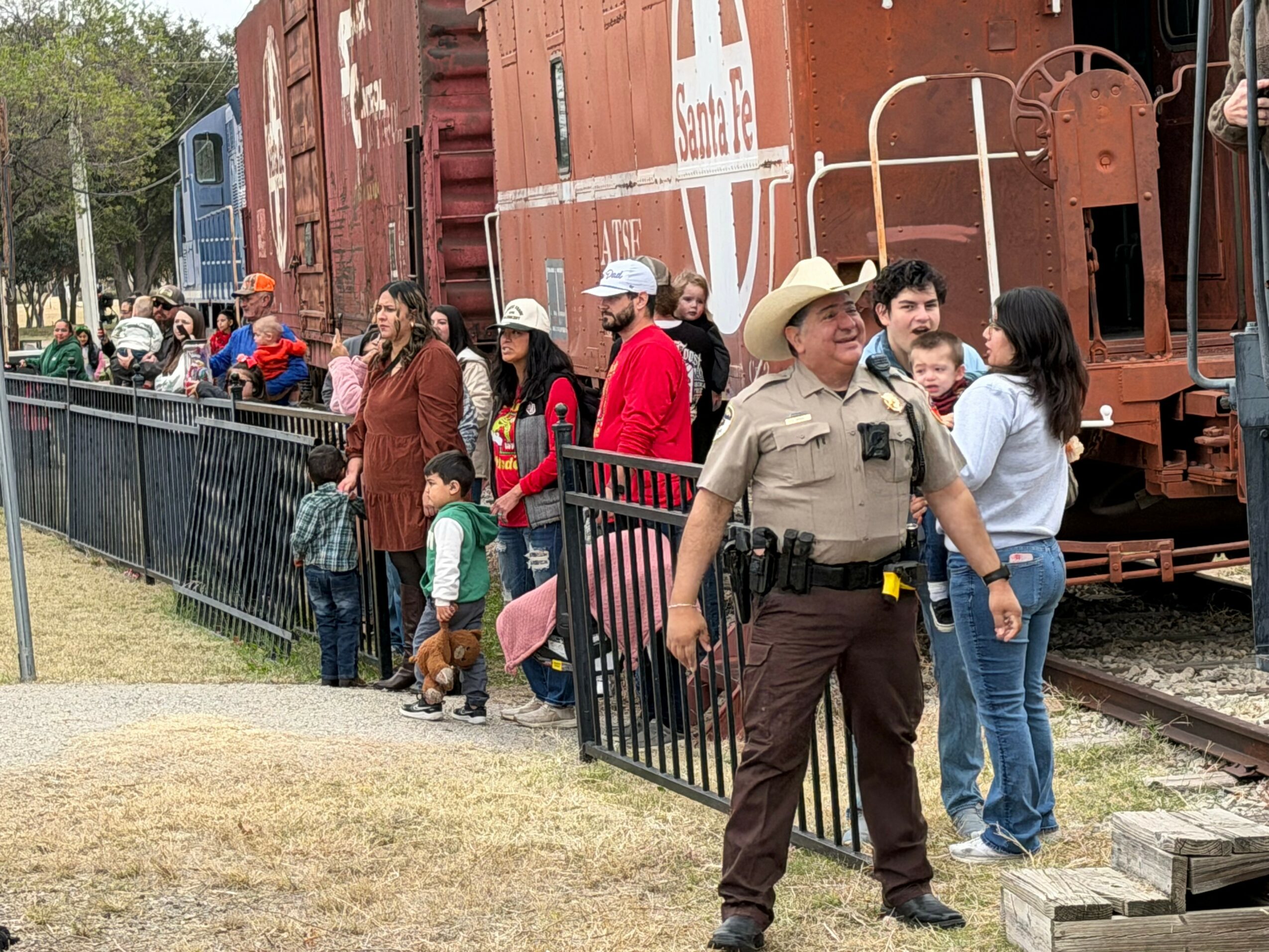 Group of people, including children, standing near a fence and train cars outdoors, some wearing hats and casual clothing.