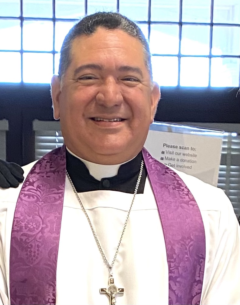 Smiling man in clerical attire with purple stole and cross necklace, standing indoors with window background.
