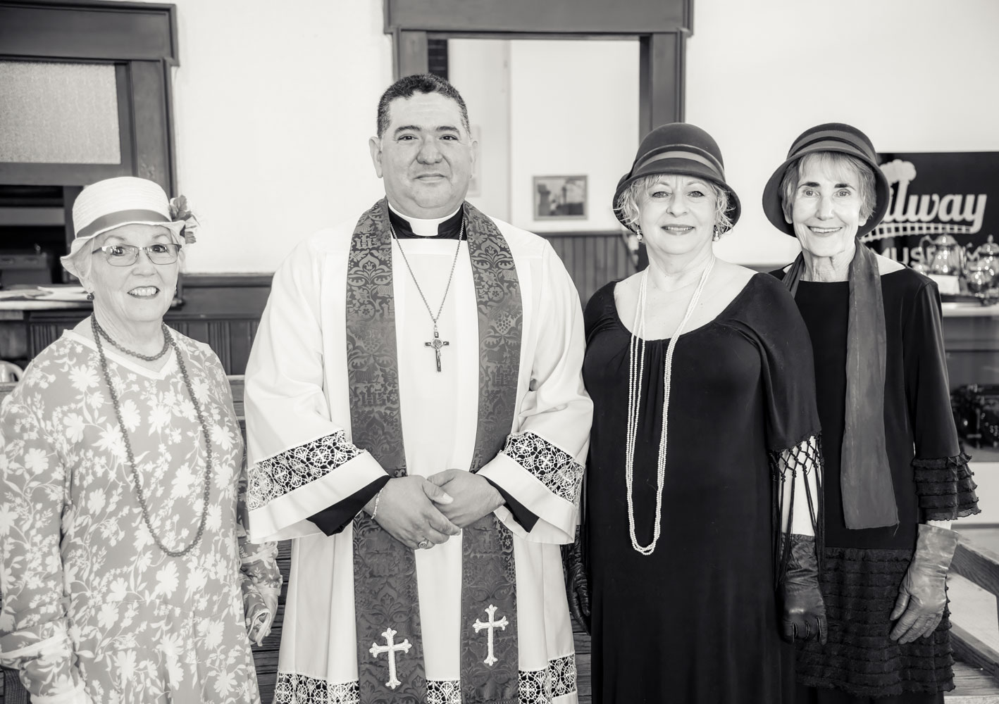 Four people dressed in vintage clothing, standing indoors, with a man in clerical attire in the center, smiling.