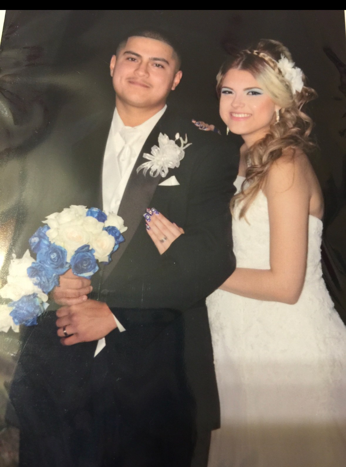 A bride and groom pose together, the groom holding a bouquet of white and blue flowers, both smiling.