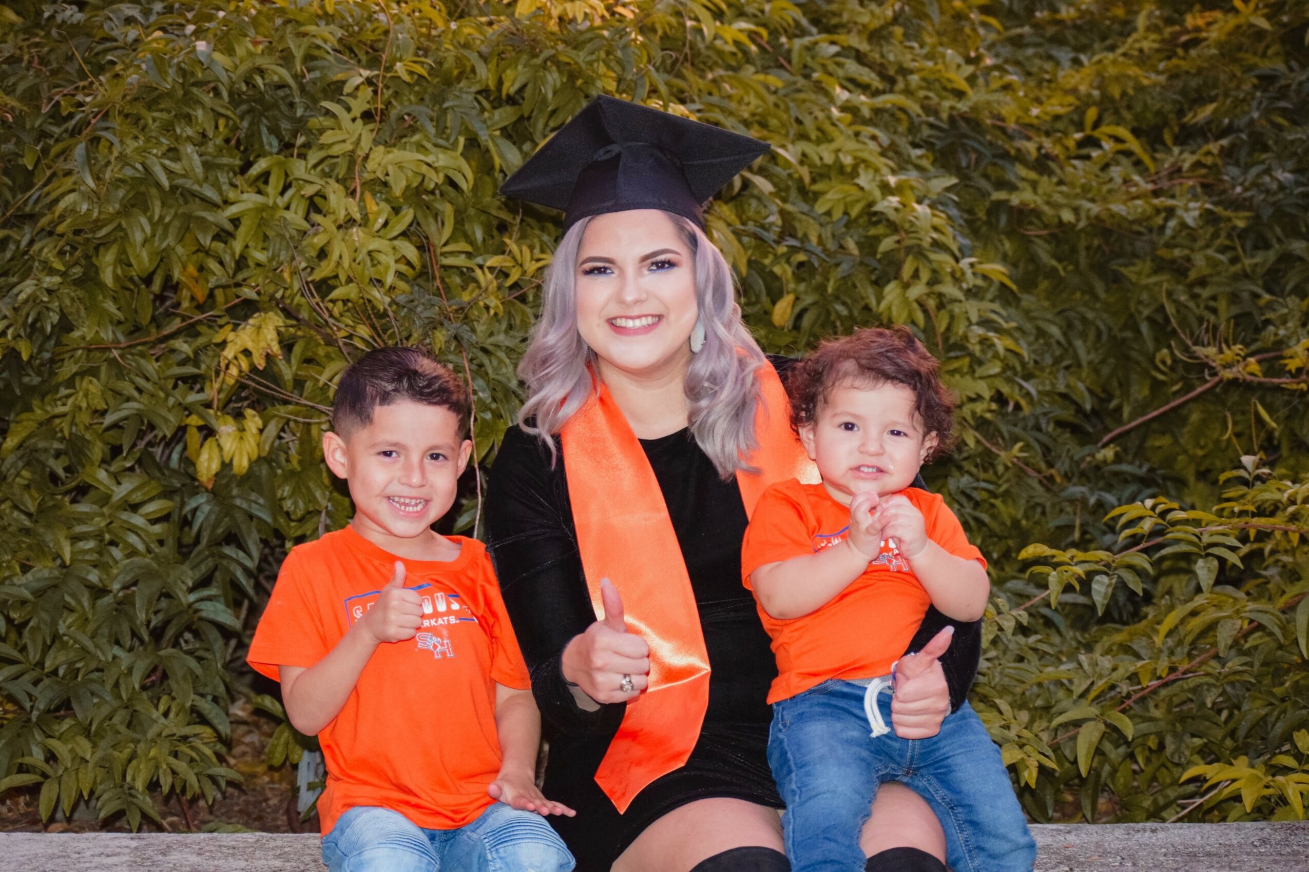 Woman in graduation cap and gown with two children outdoors, all smiling and giving thumbs up, surrounded by green foliage.