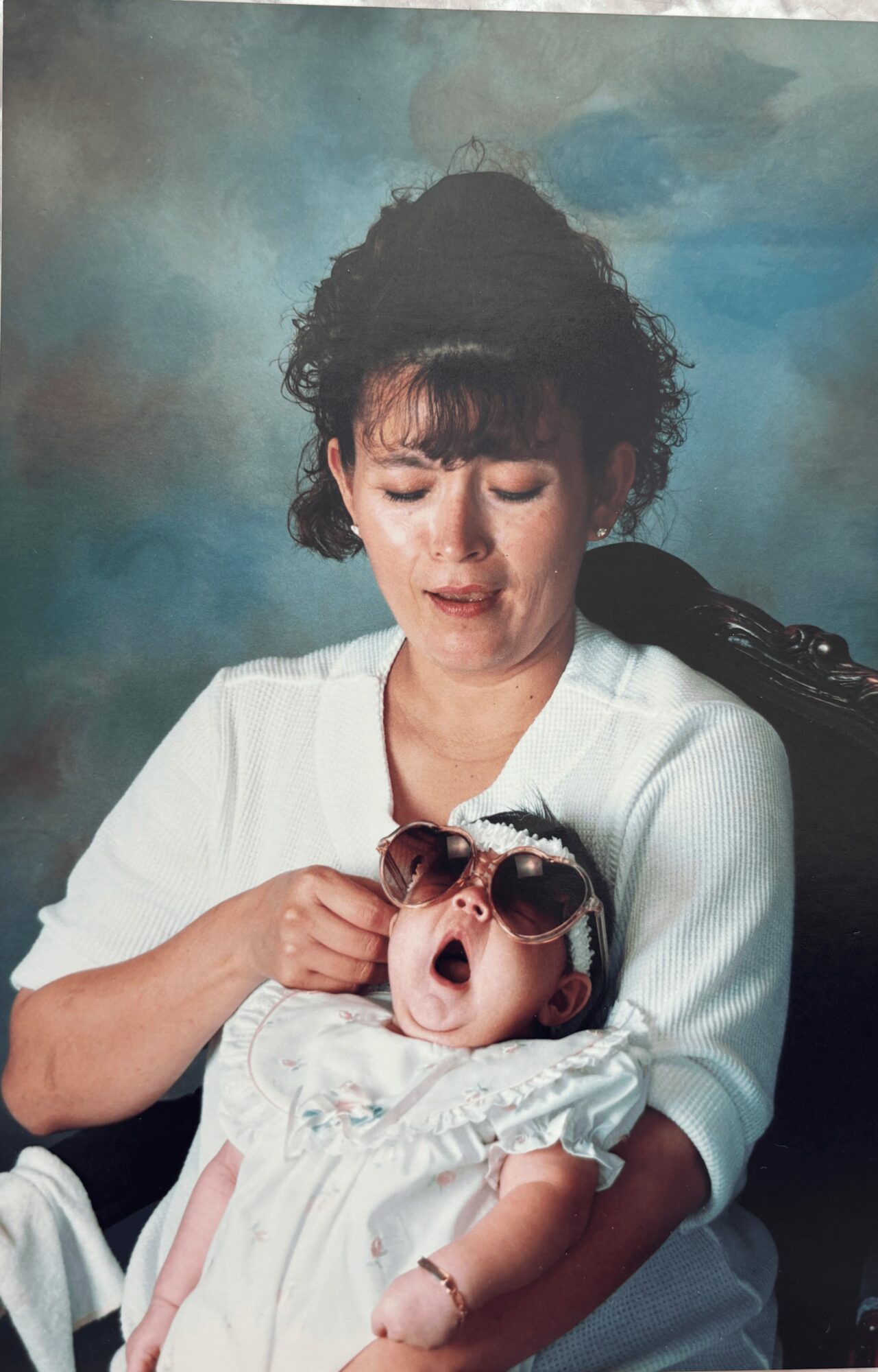 Woman holding a baby girl wearing sunglasses, both smiling, against a blue background.