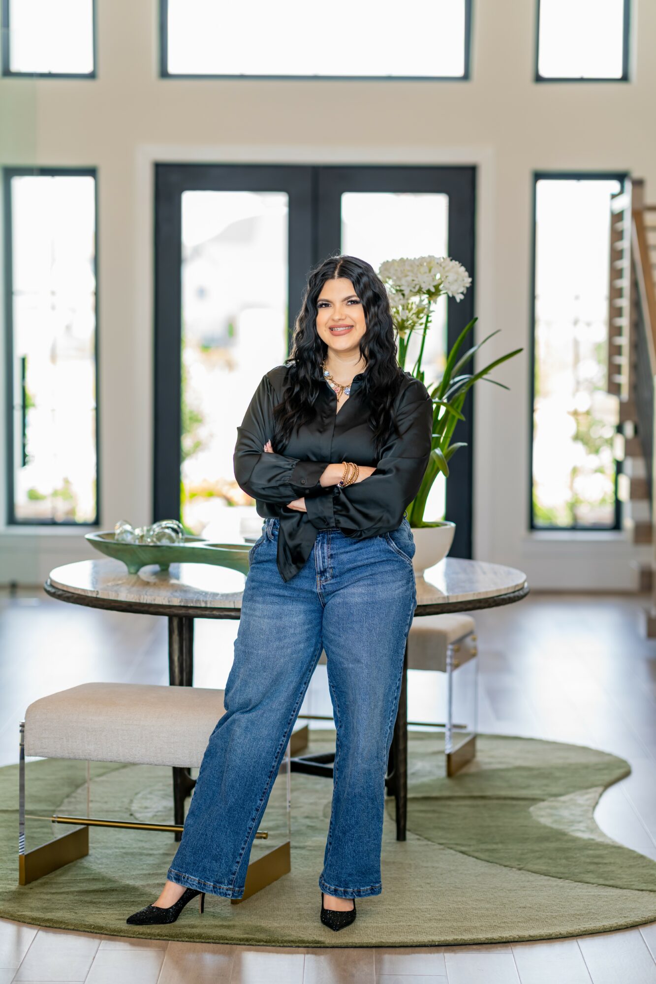 Woman with long dark hair in black top and blue jeans standing in a bright room with large windows and a round table.