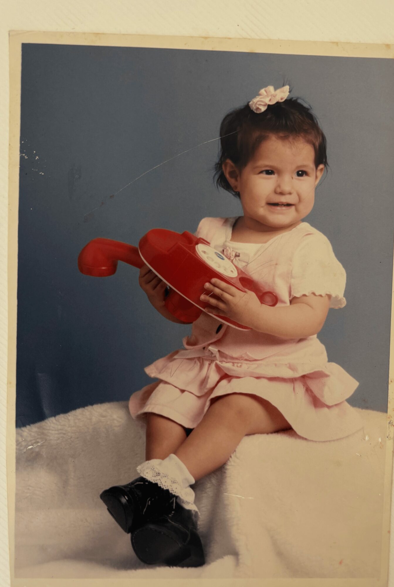 Young girl sitting on a white surface holding a red toy guitar, wearing a light pink dress and a hair bow.