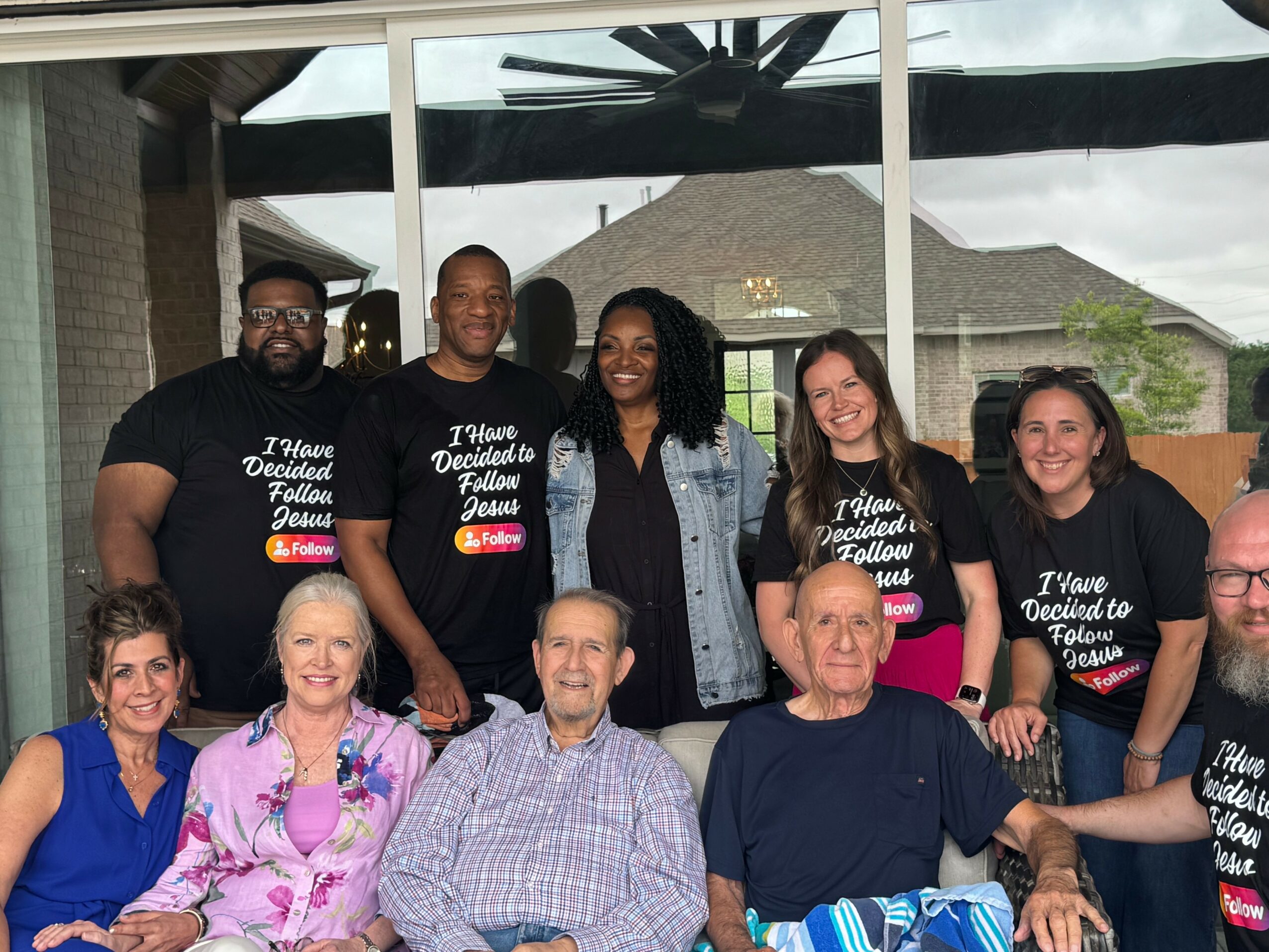 Group of eight people smiling outdoors, some wearing matching black T-shirts, in front of a glass window and patio area.