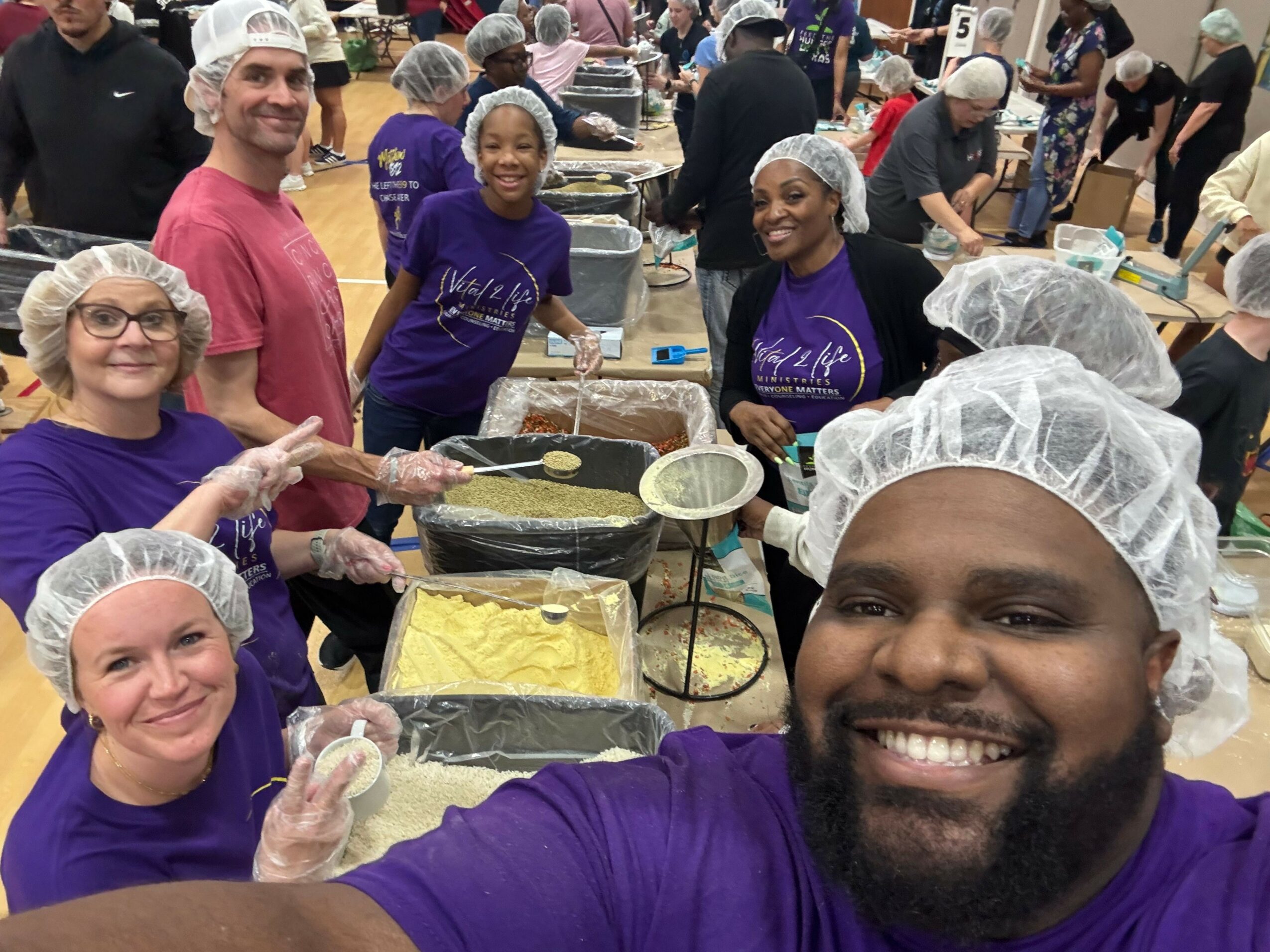 Group of people wearing hairnets and aprons, preparing food together in a kitchen, smiling and working around a table.