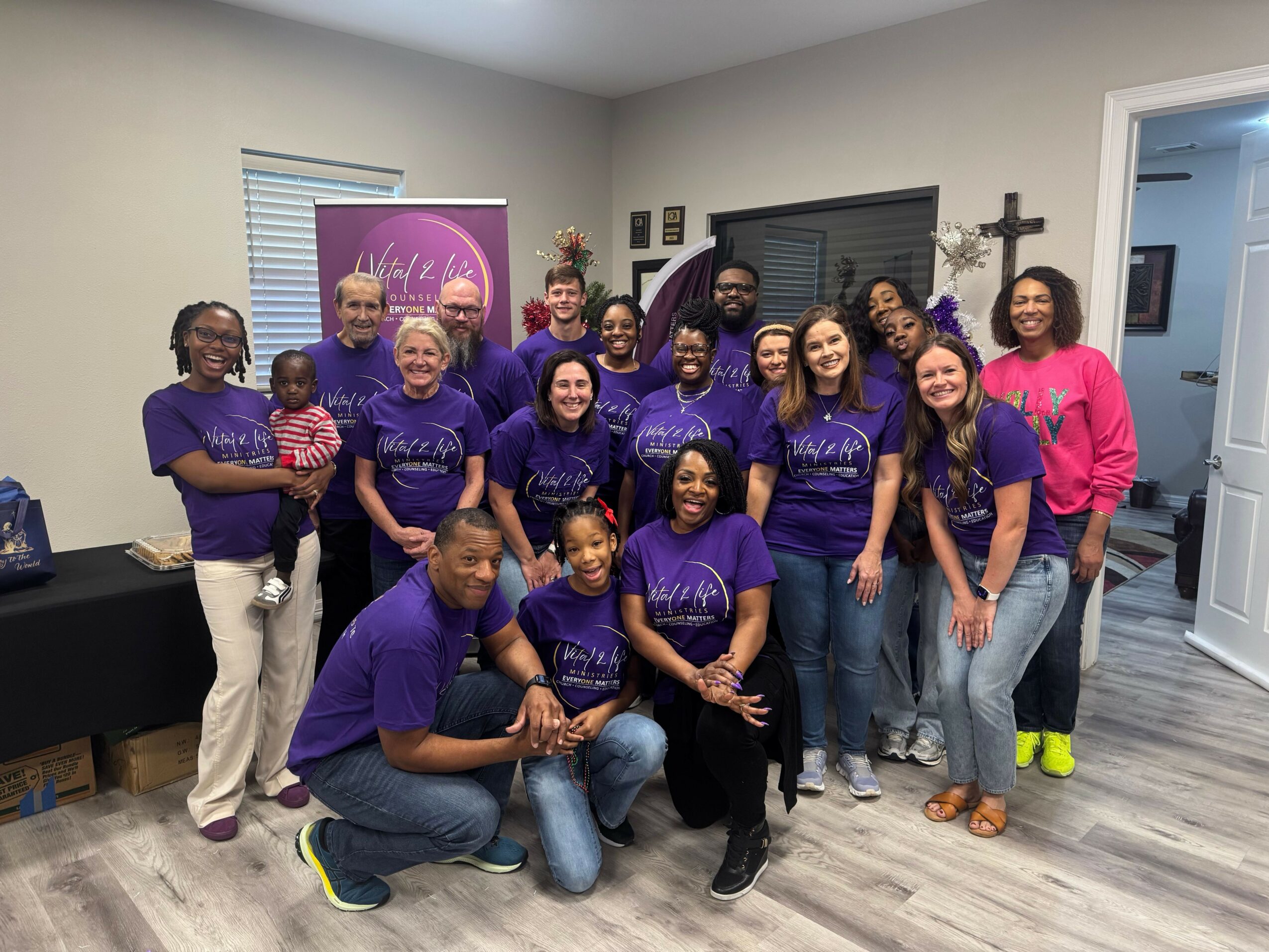 Group of people sitting on the floor, smiling, wearing purple shirts, in a room with white walls and windows.