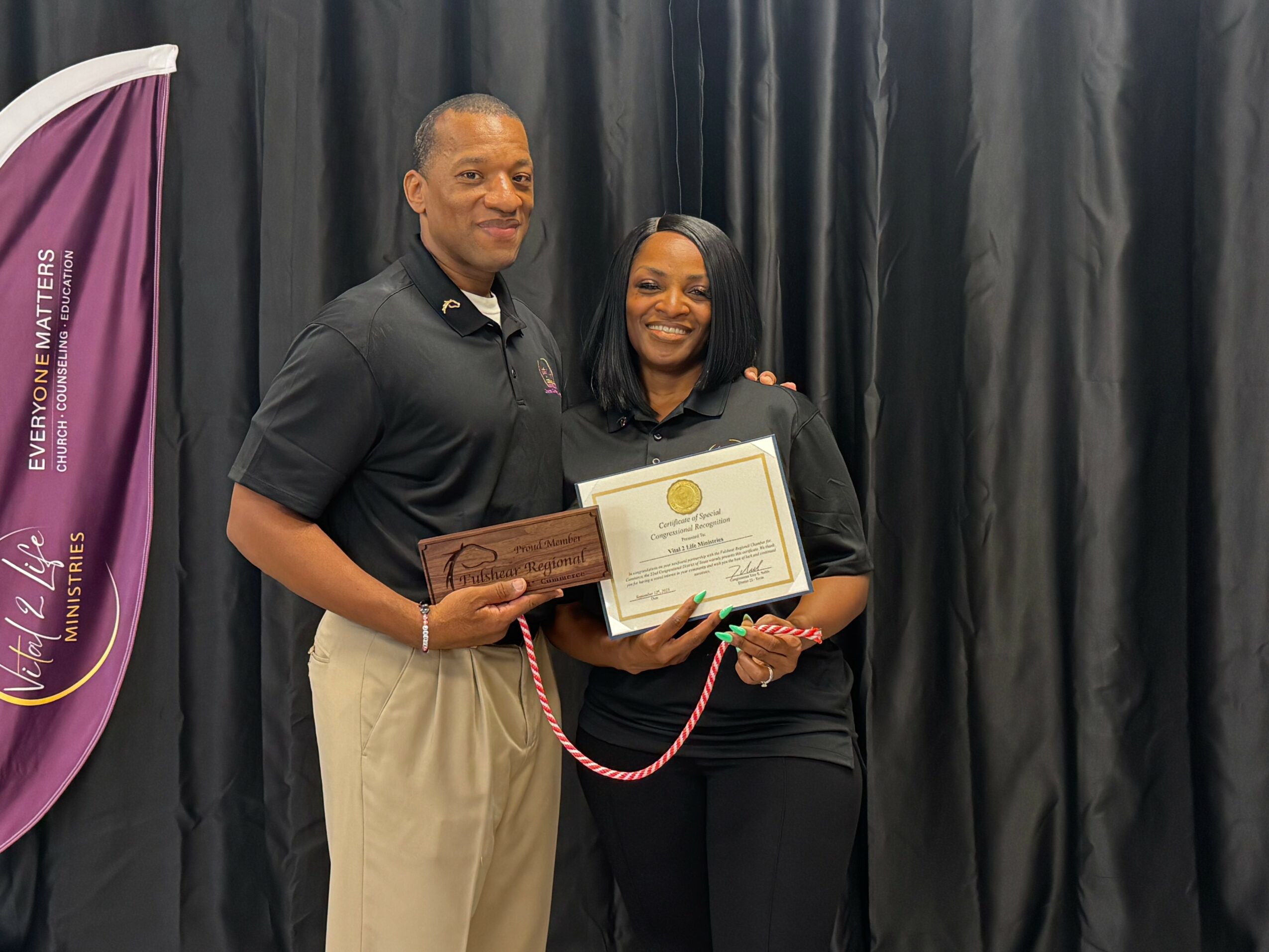 Two people standing in front of black curtains, holding a certificate and a plaque, smiling at the camera.