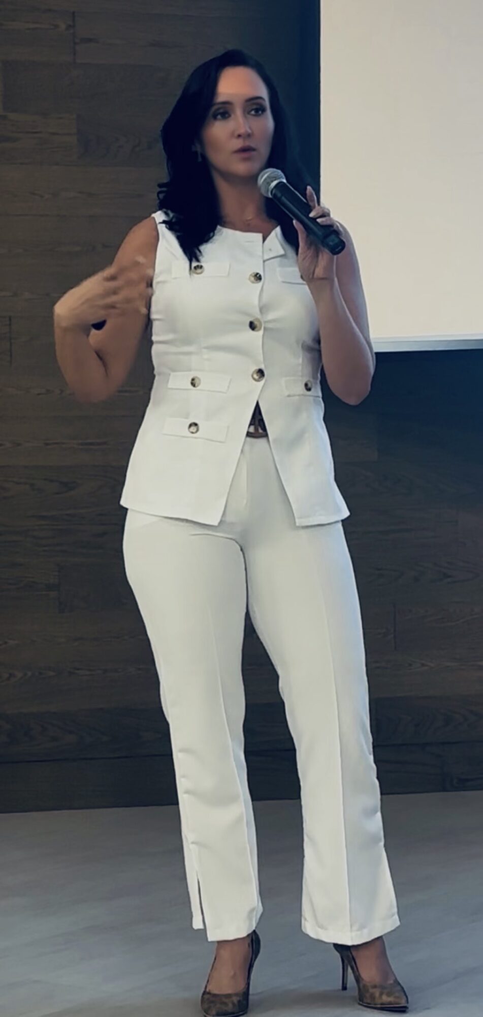 Woman in white suit holding microphone, speaking in front of a screen and wooden wall.