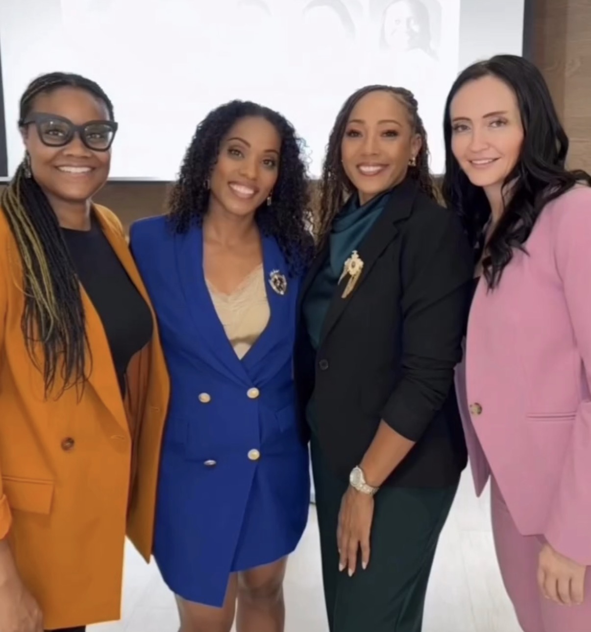 Four women standing together, smiling, dressed in colorful blazers, in an indoor setting.