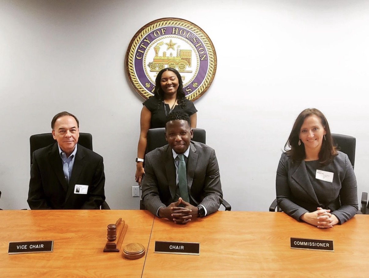 Four people seated and standing at a conference table with a seal on the wall behind them.