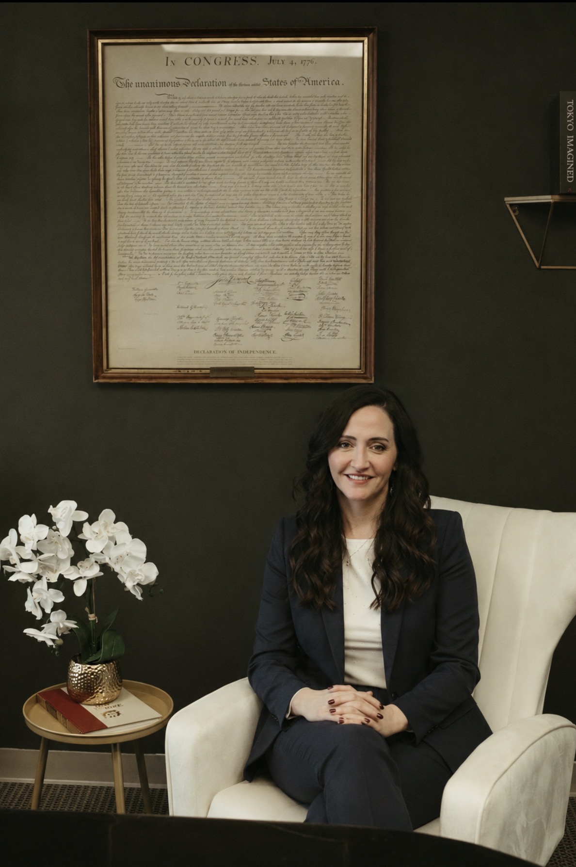 Woman with long dark hair sitting in a white armchair, smiling, in a room with dark green wall and framed document.