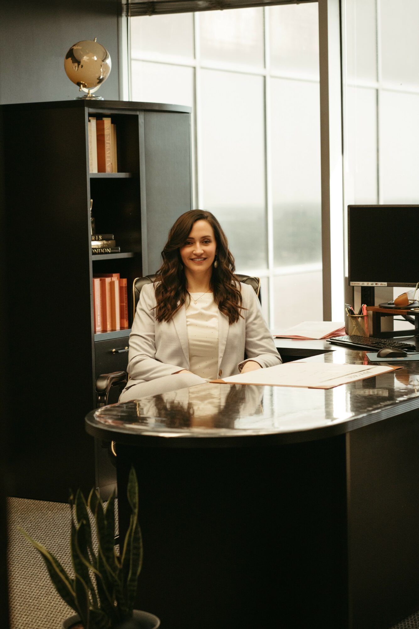 Woman sitting at a desk in an office with large windows, bookshelves, and a computer.