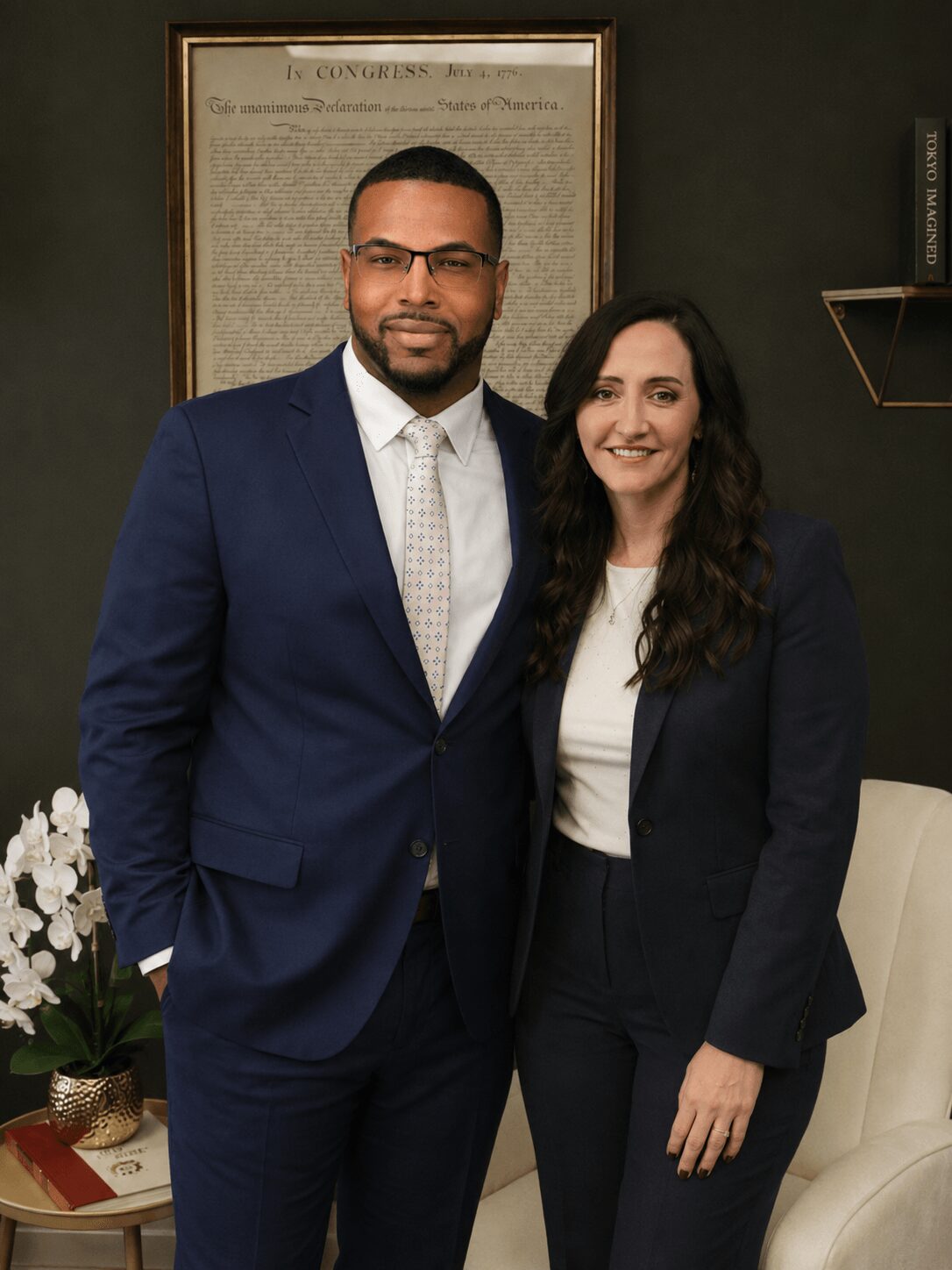Two people, a man in a suit and a woman in a blazer, stand together indoors, smiling at the camera.