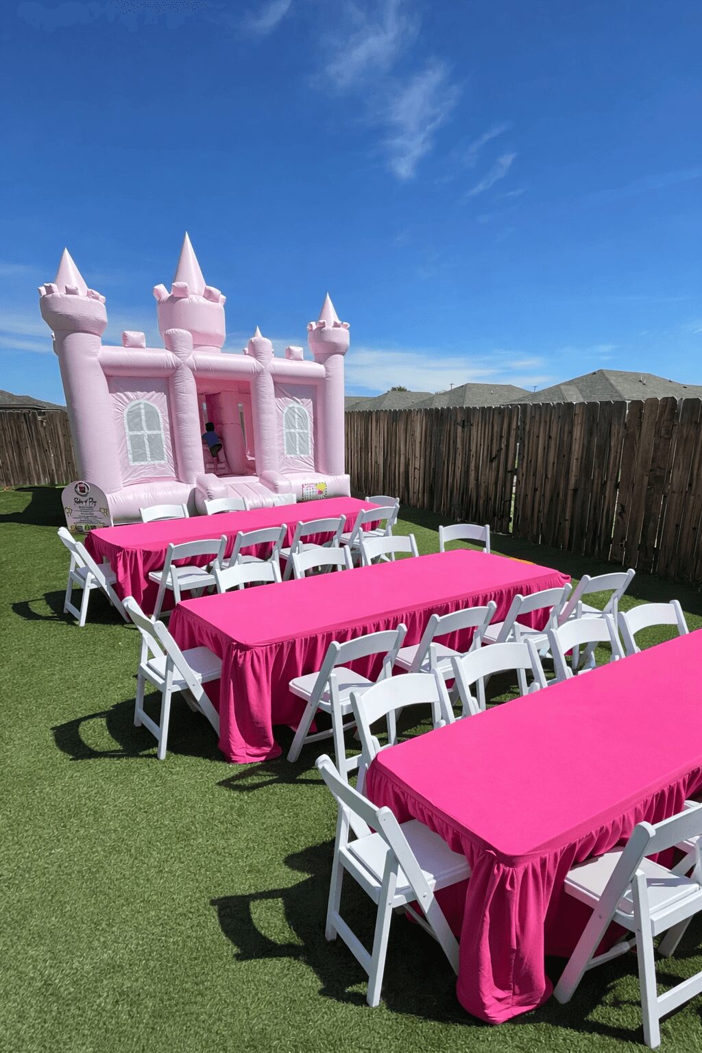 Pink castle-themed inflatable bounce house with white towers, set outdoors with tables covered in pink cloths and white chairs, on grass.