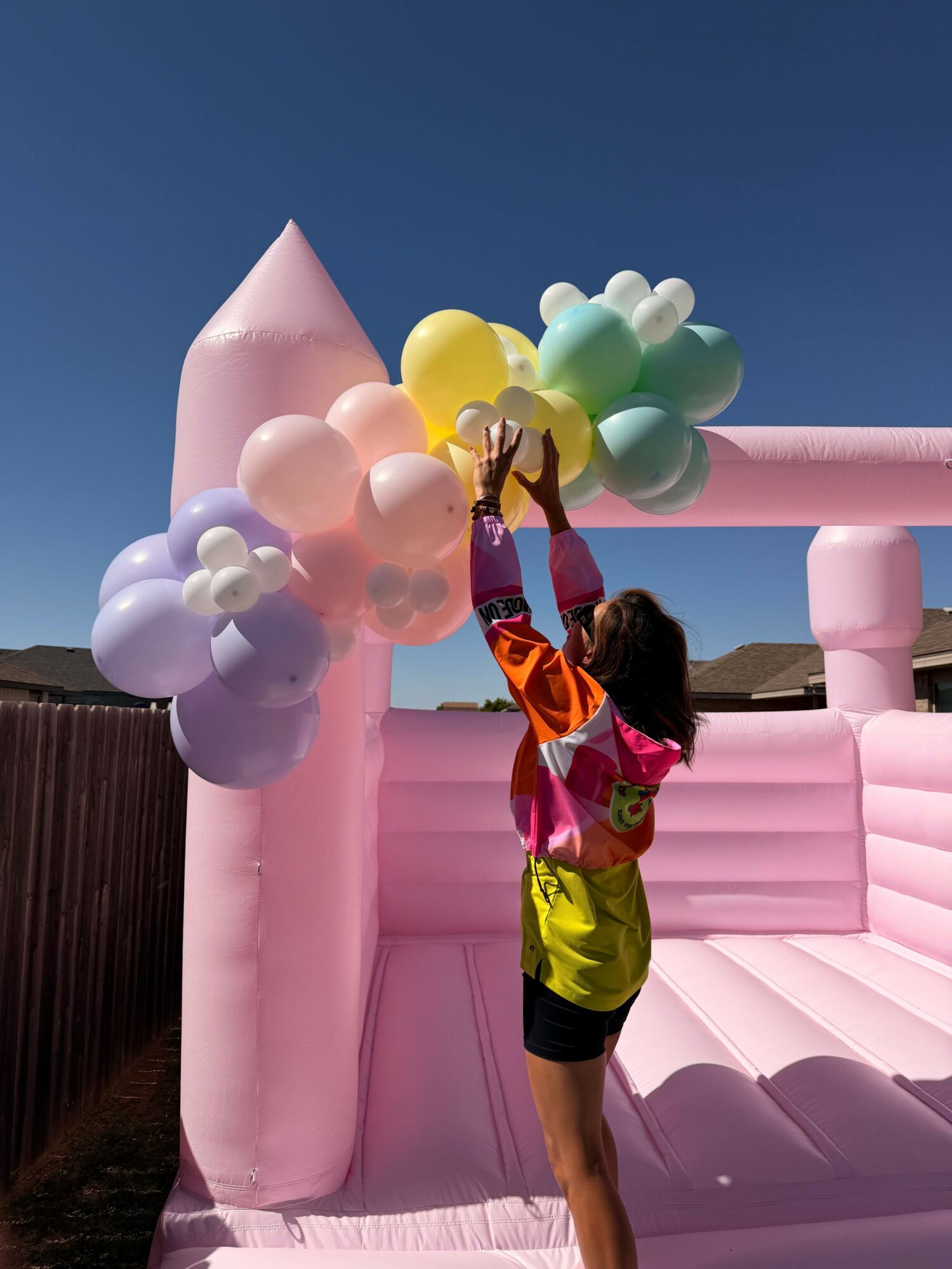Child holding colorful balloons on pink inflatable structure outdoors under blue sky.