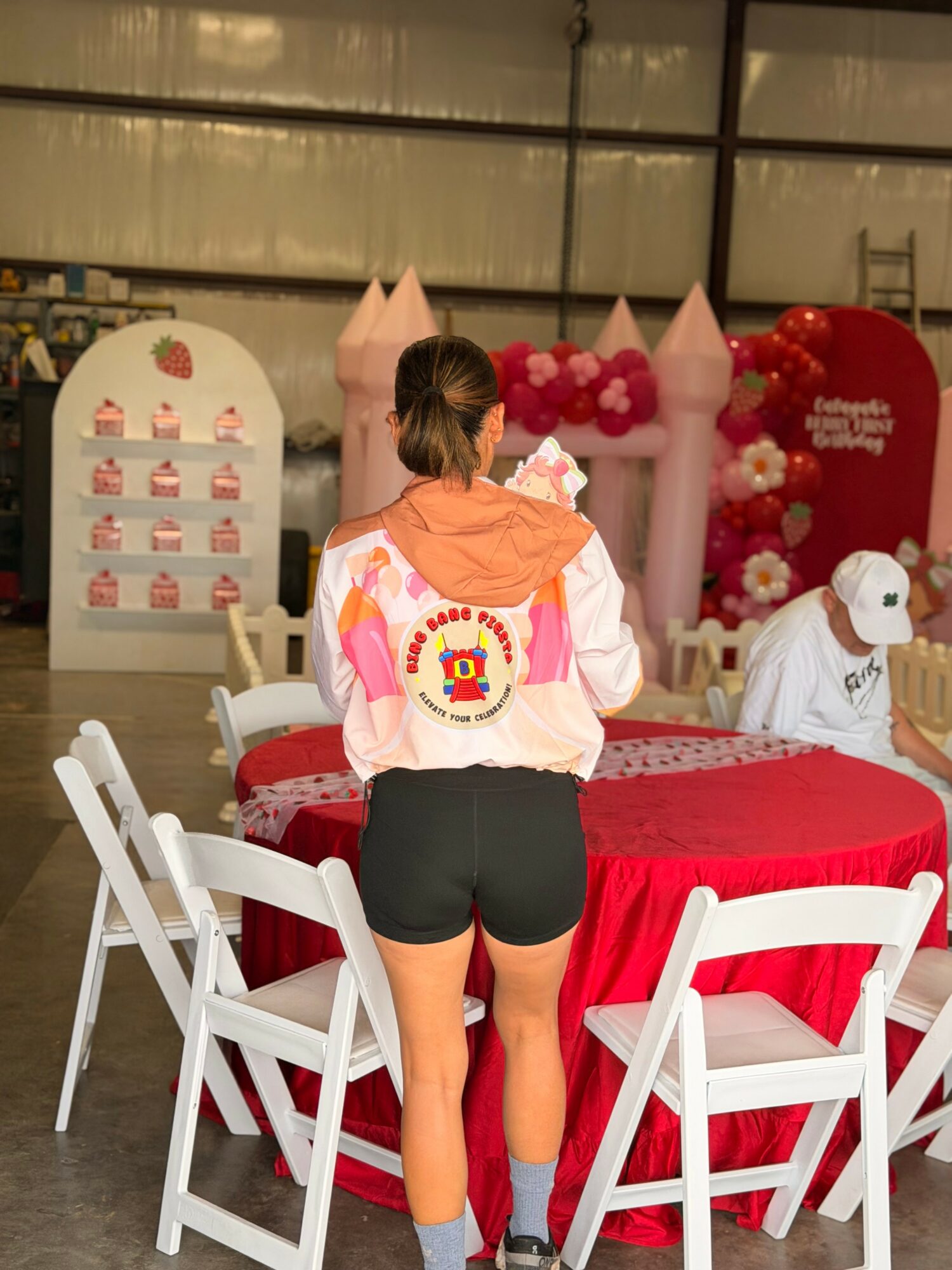 Person with a colorful jacket and black shorts standing near a decorated table with pink and red balloons, chairs, and a cake, in a large indoor space.