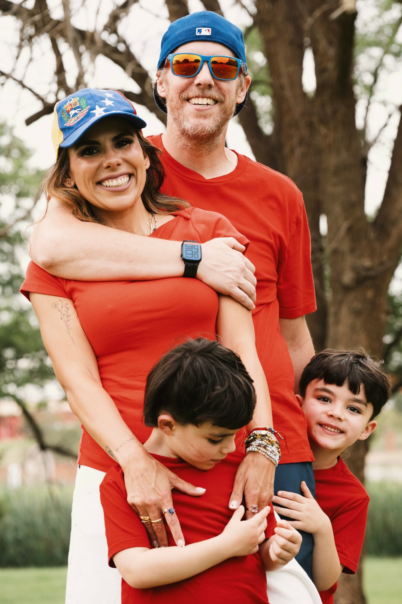 A smiling family of four outdoors, with two children and two adults, all wearing red shirts and hats, standing near trees.