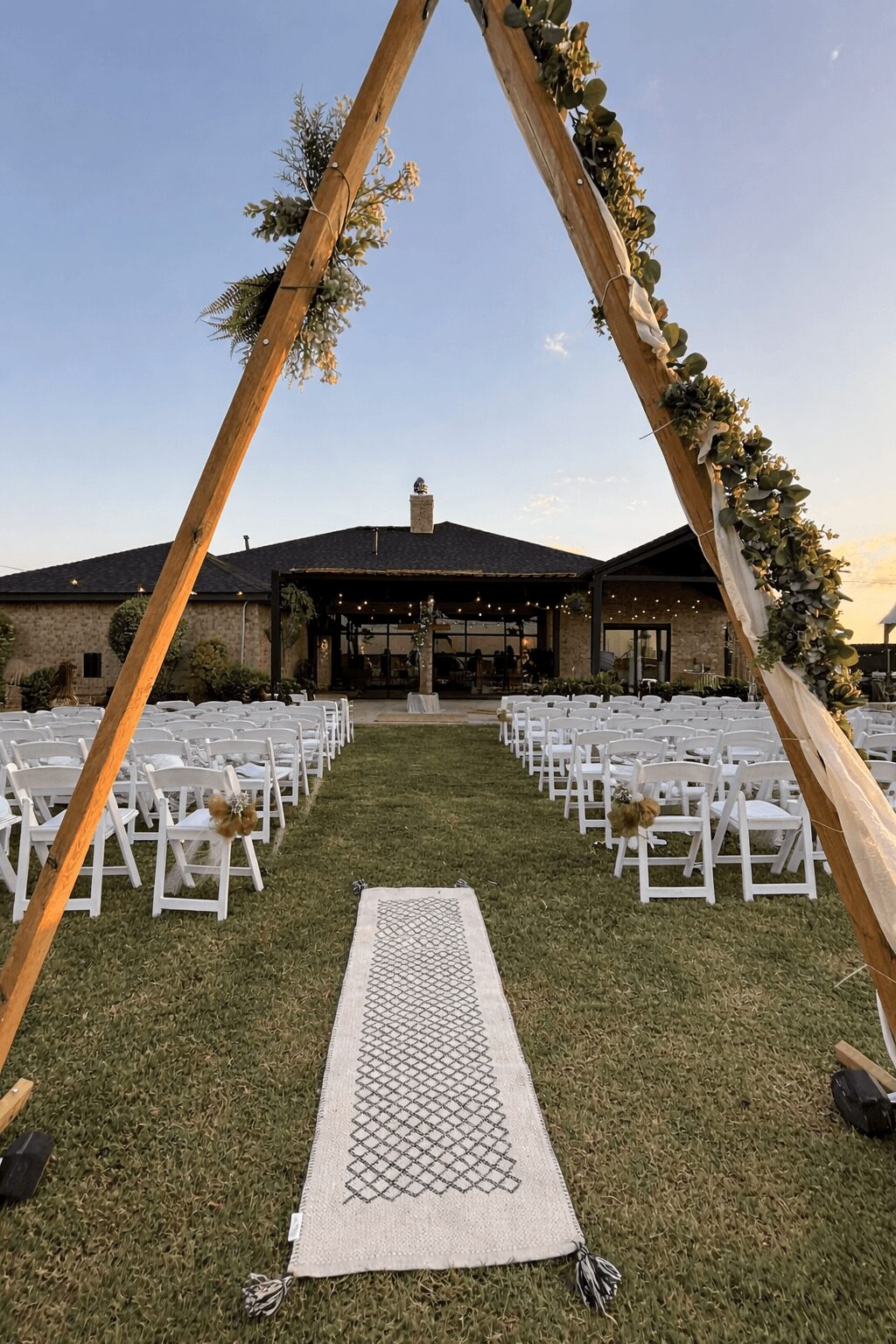 Outdoor wedding setup with chairs arranged on grass, decorated arch, and building in background, under clear sky.