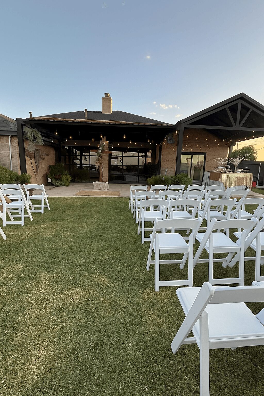 Outdoor event space with white chairs on grass, a covered pavilion, string lights, and a building in the background.