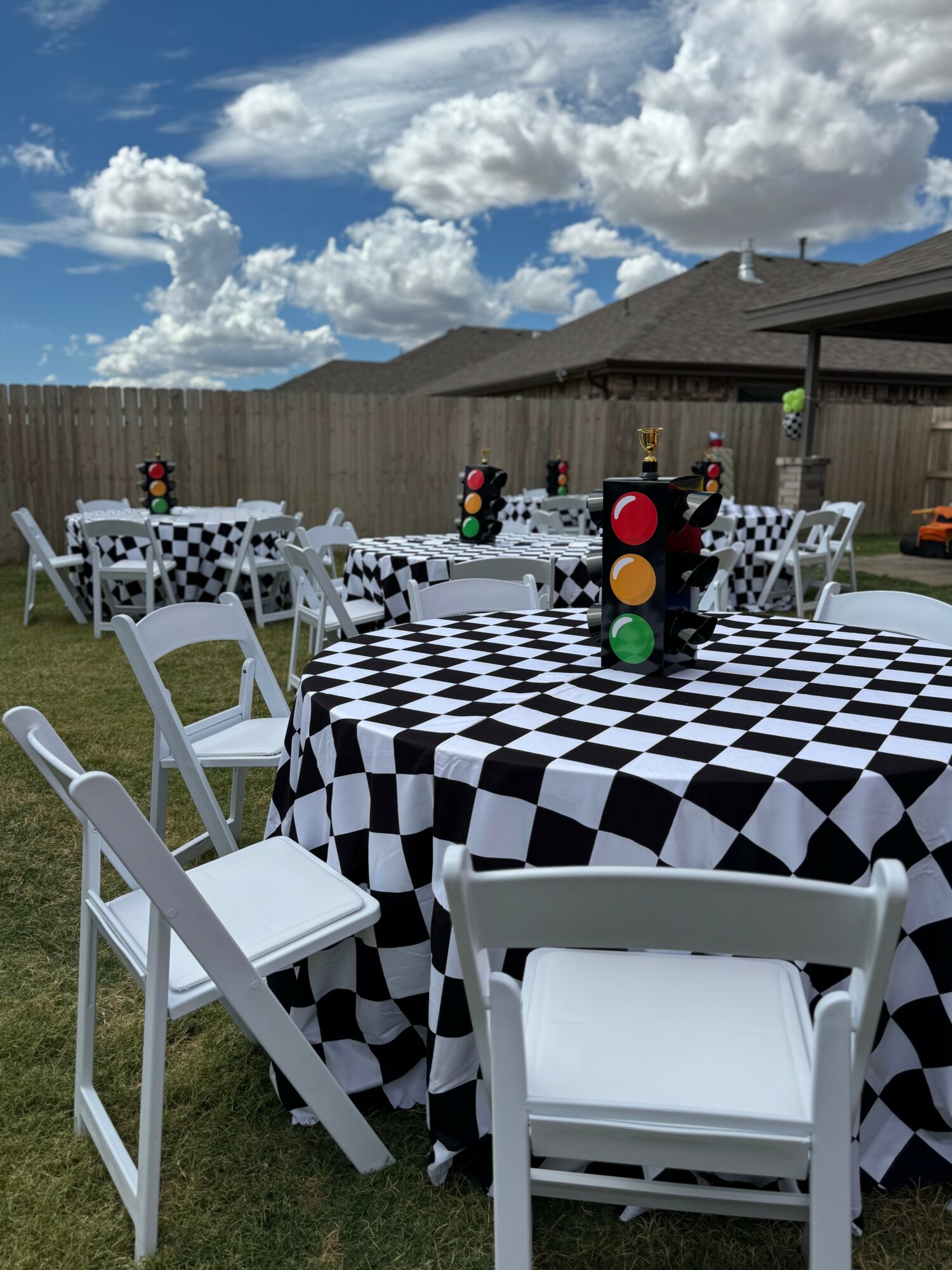 Outdoor party setup with tables covered in black and white checkered tablecloths, white chairs, and traffic light decorations, under a partly cloudy sky.