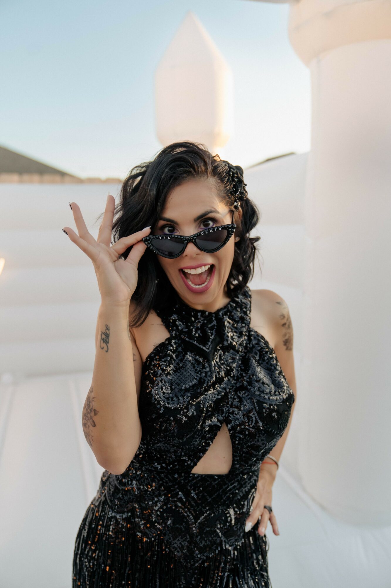 Woman with dark hair wearing sunglasses and a black dress, smiling and holding sunglasses, outdoors with white architectural elements.