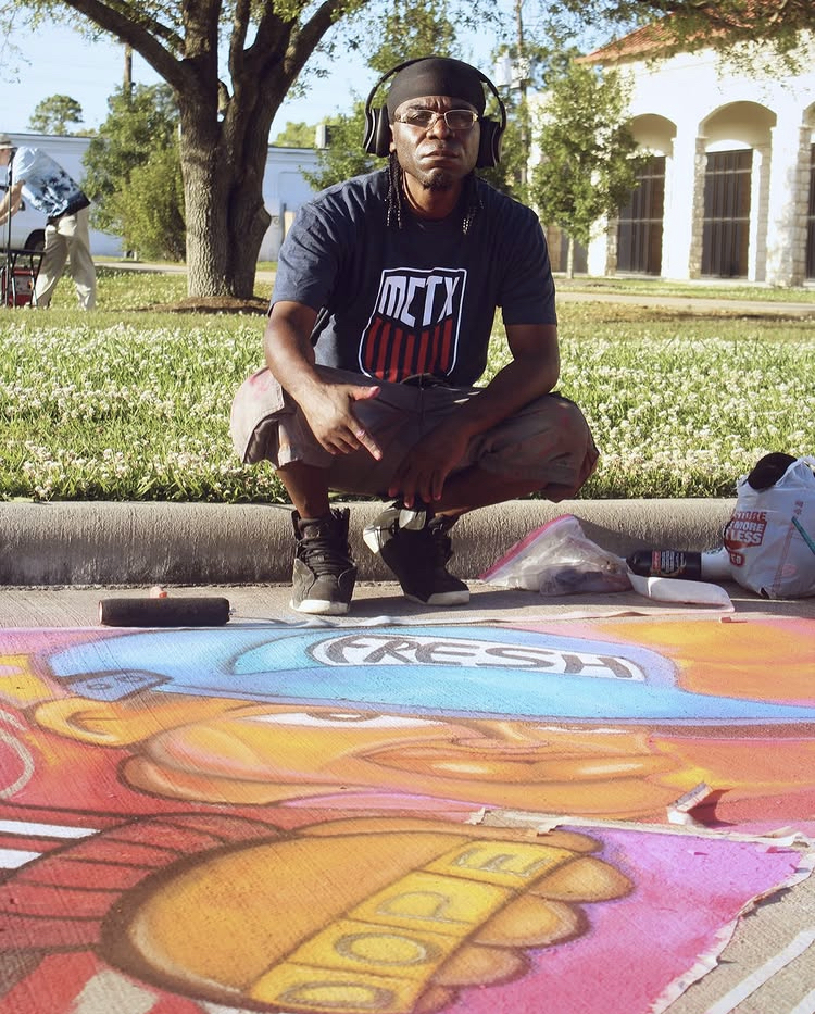 Person sitting cross-legged on sidewalk with headphones, wearing a black T-shirt, in front of colorful chalk art on pavement.