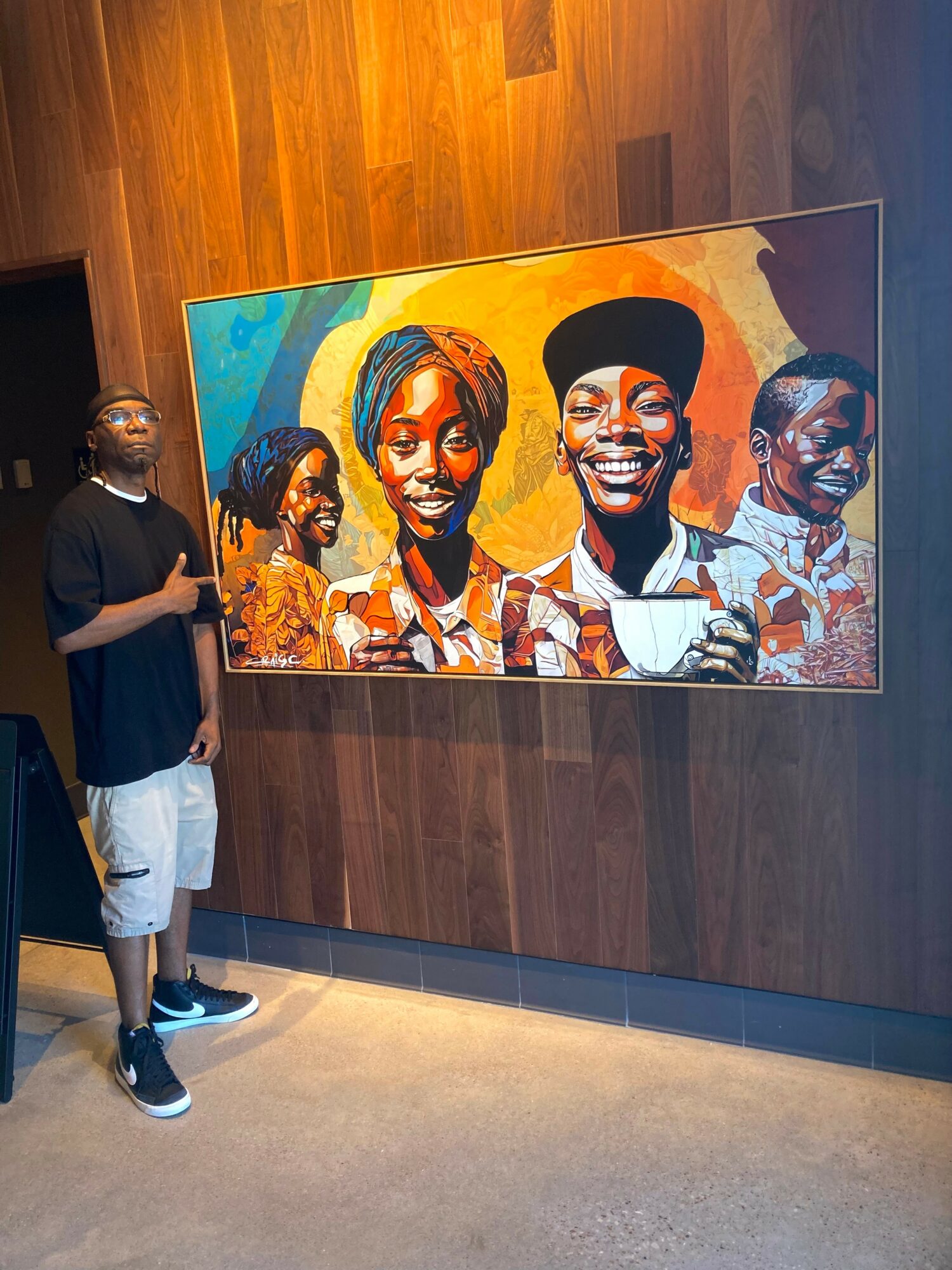Person standing next to colorful portrait painting on wooden wall, smiling, wearing black shirt and shorts.