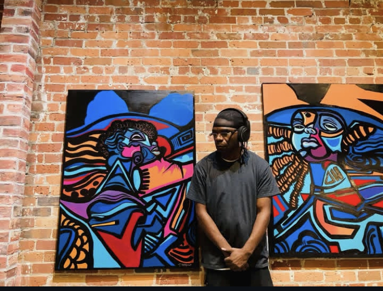 Person with glasses and black shirt stands in front of colorful abstract paintings on brick wall.