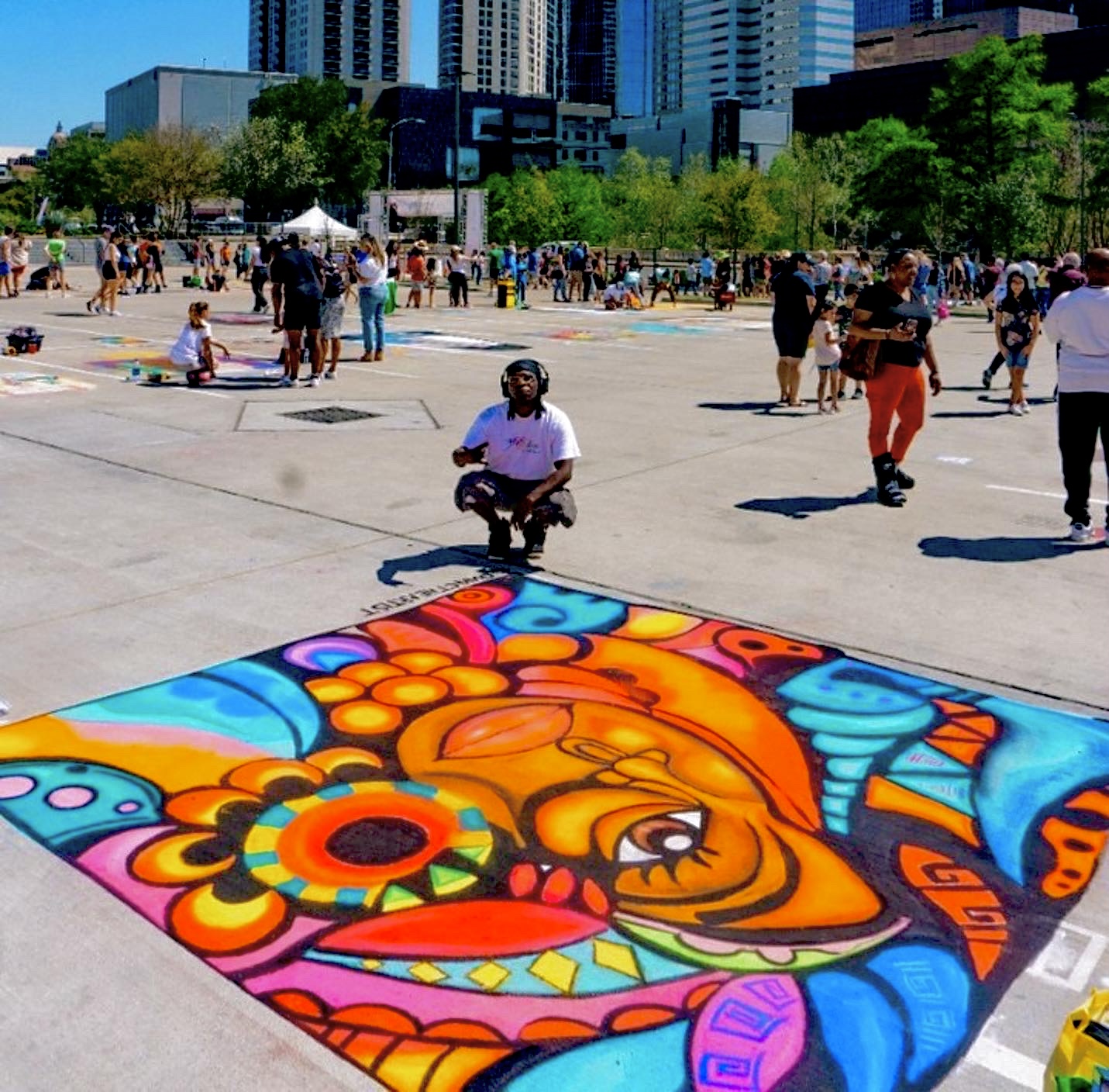 Colorful chalk art on pavement with people in an outdoor urban space, buildings, and trees in background.