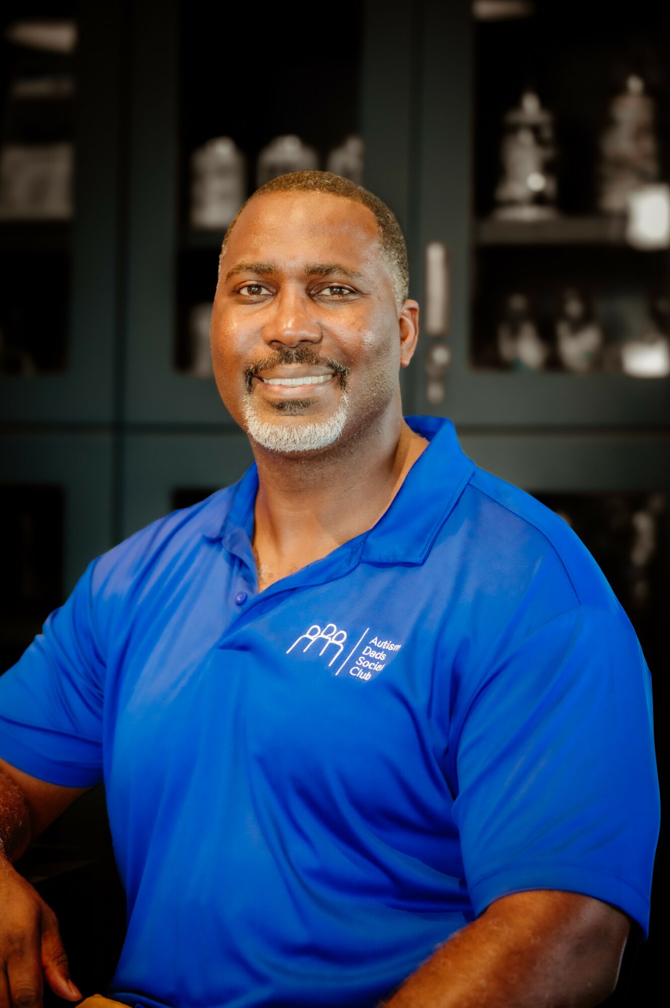 Man with short hair and beard wearing a blue polo shirt, smiling, in front of shelves with decorative items.